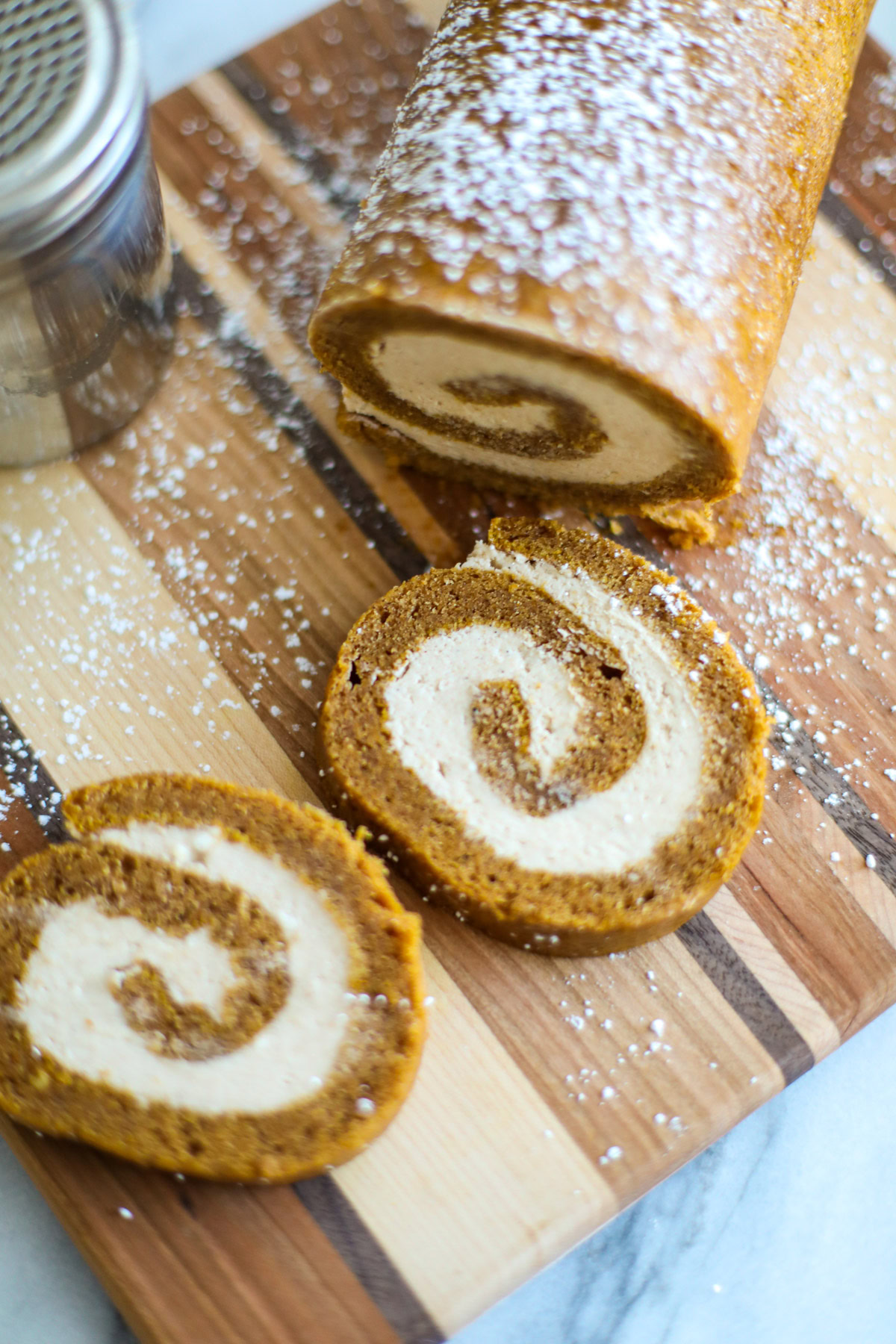 A sliced pumpkin roll siting on a striped cutting board with a silver powdered sugar shaker.