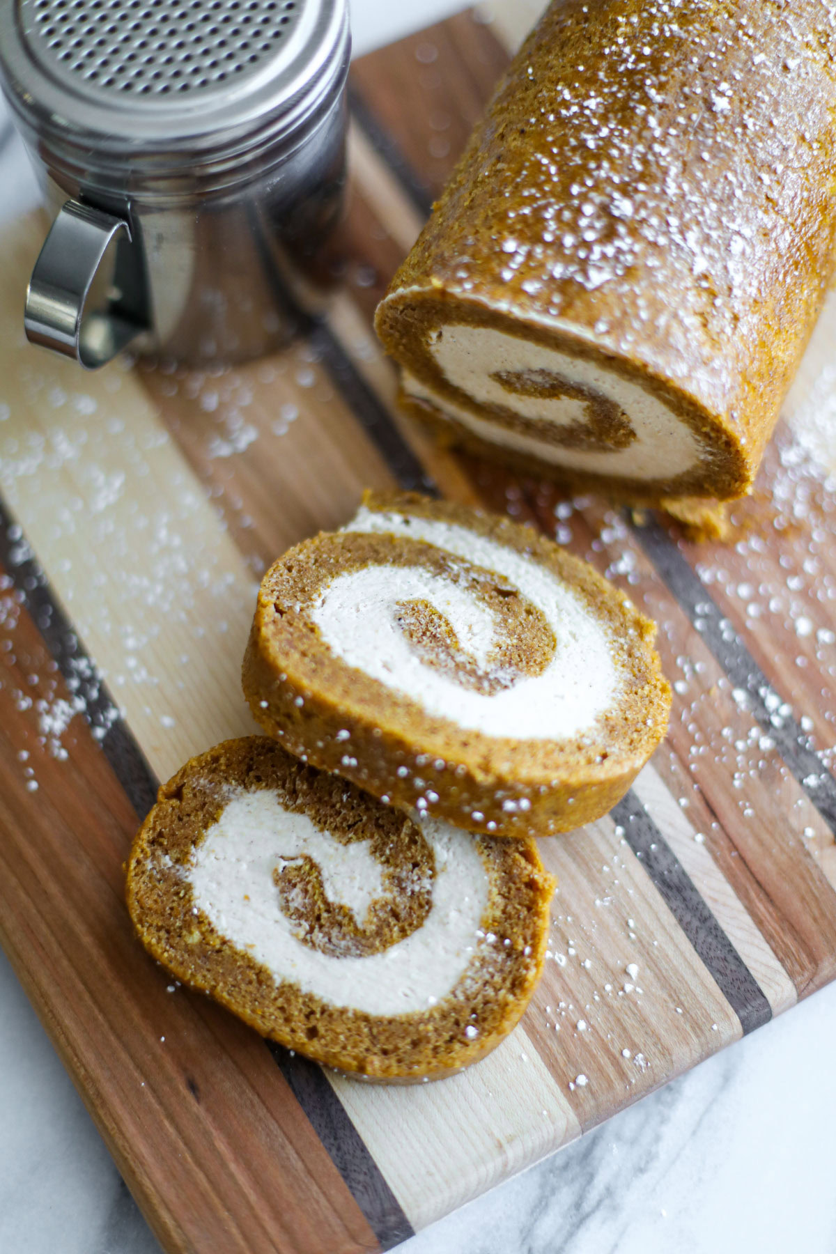 A sliced pumpkin roll showing the cream cheese swirl, with a powdered sugar shaker.