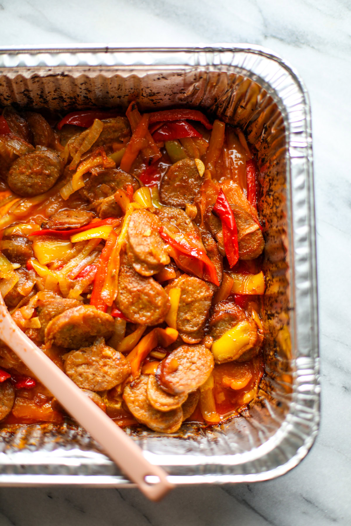 The corner of a rectangular aluminum pan with crispy sausage slices and caramelized onions and peppers with a pink serving spoon.