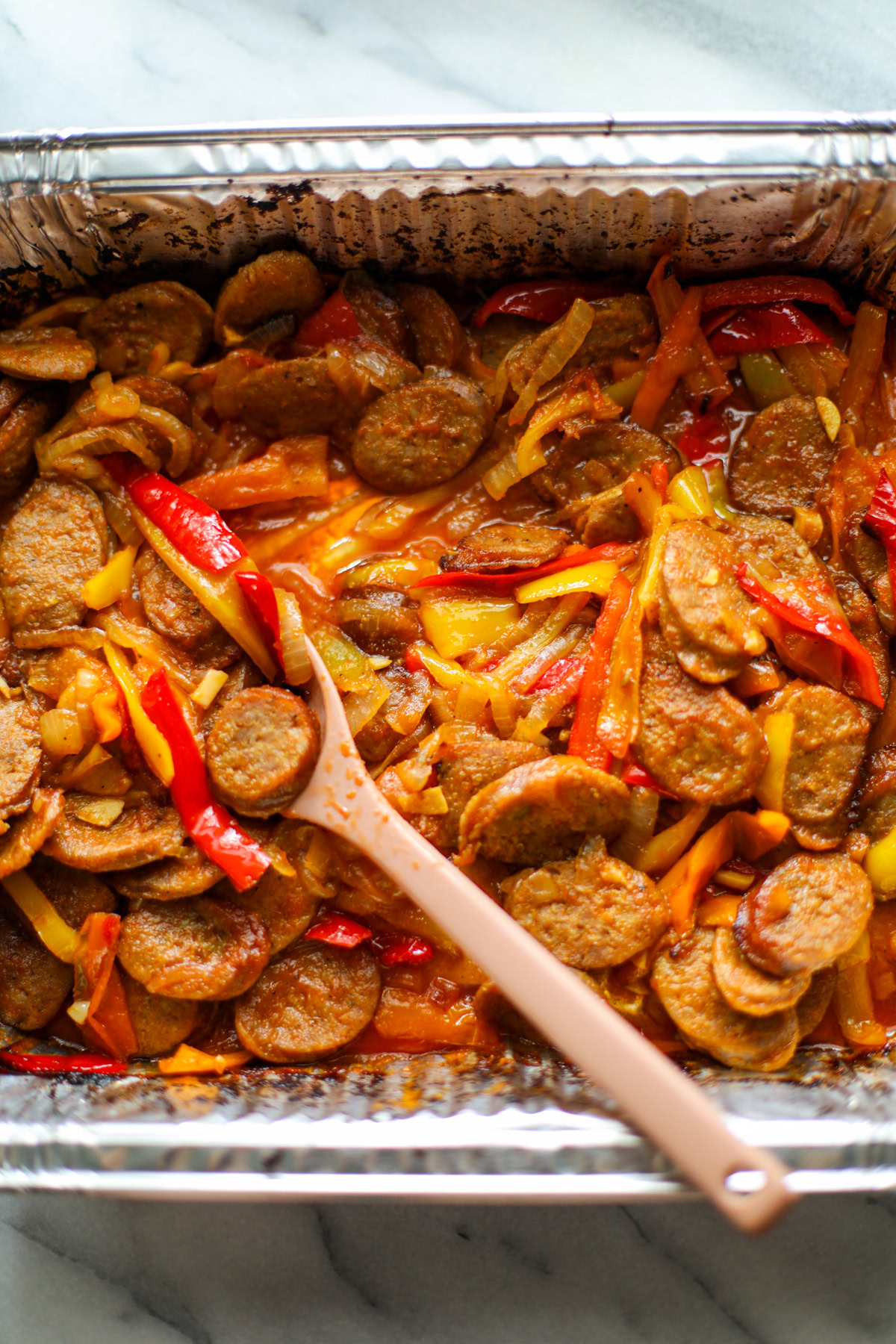 A rectangular aluminum pan with crispy sausage slices and caramelized onions and peppers with a pink serving spoon.