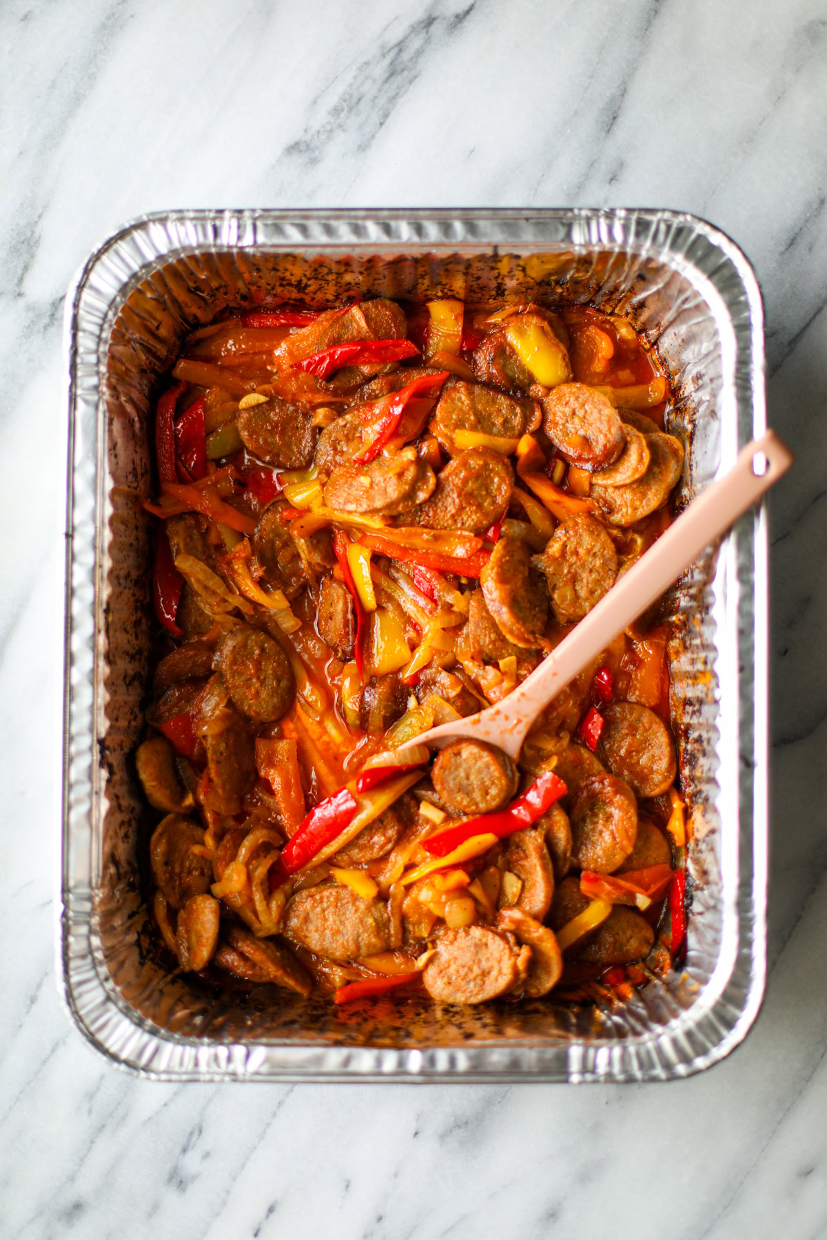 A rectangular aluminum pan with crispy sausage slices and caramelized onions and peppers with a pink serving spoon.