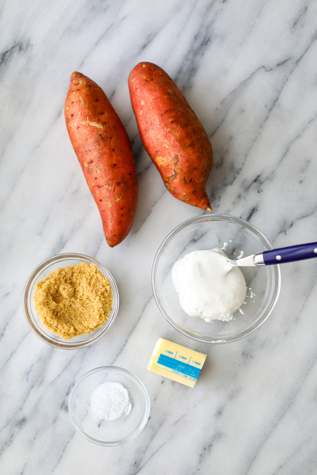 Two sweet potatoes, a bowl of sugar, a bowl of marshmallow fluff with a blue spoon, 3 tablespoons of butter, and a small bowl of salt.