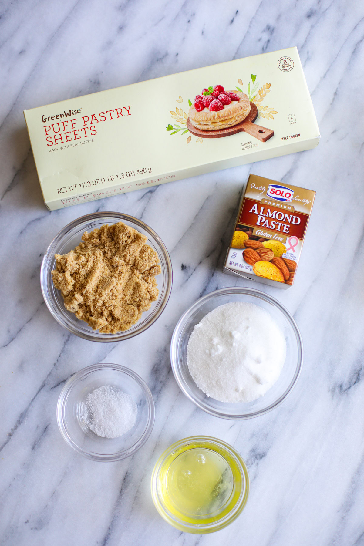 A box of puff pastry sheets, a box of almond paste, a glass bowl of light brown sugar, a glass bowls of white sugar, and two small glass bowls filled with egg white and salt.