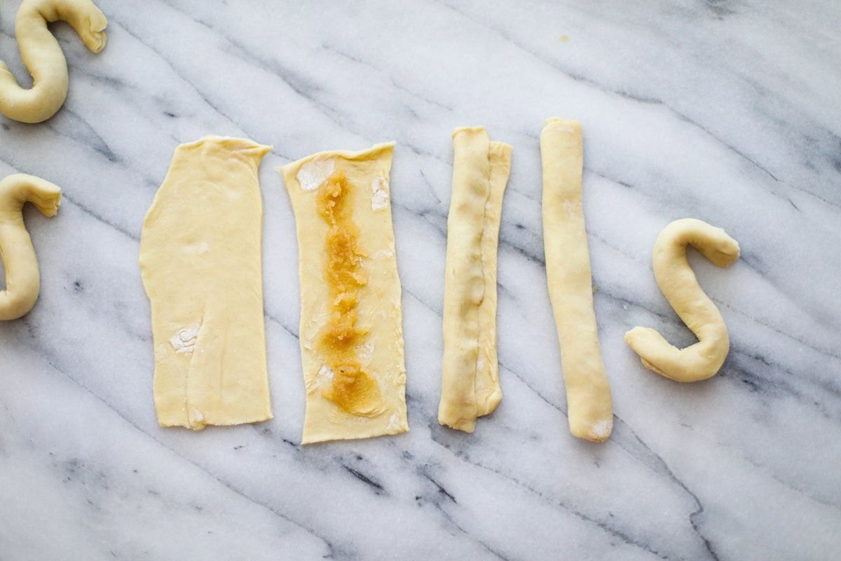 A piece of puff pastry with a spoonful of almond paste, rolled into a log, and then shaped into an S on a marble board. 