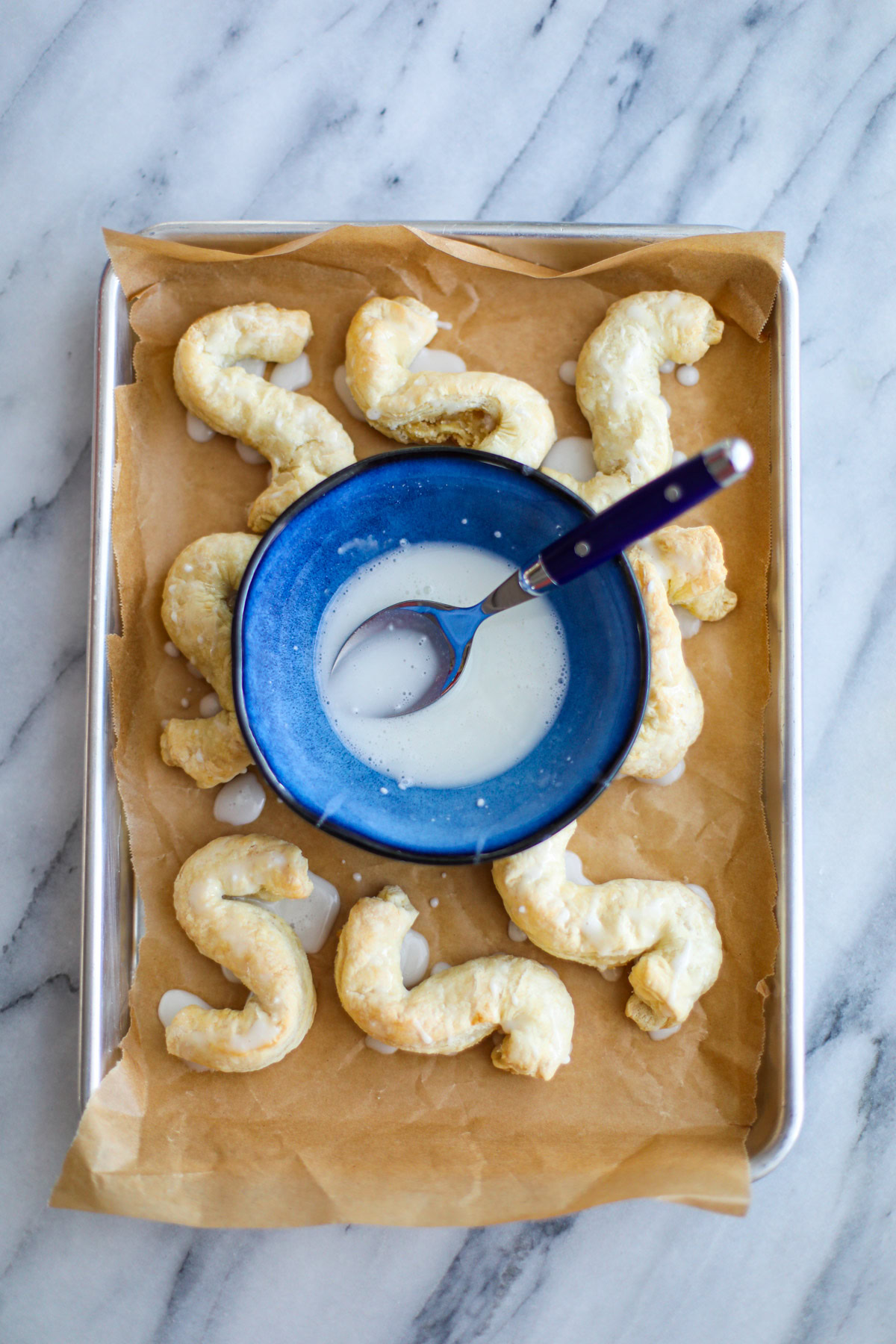 A blue bowl filled with glaze with a blue spoon sitting atop a baking sheet of almond S cookies.