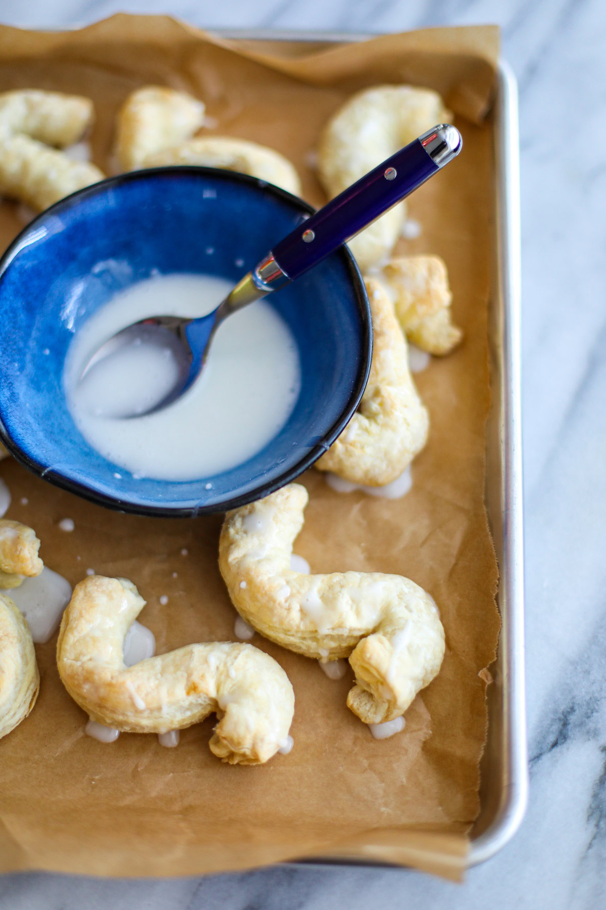 A blue bowl filled with glaze with a blue spoon sitting atop a baking sheet of almond S cookies.
