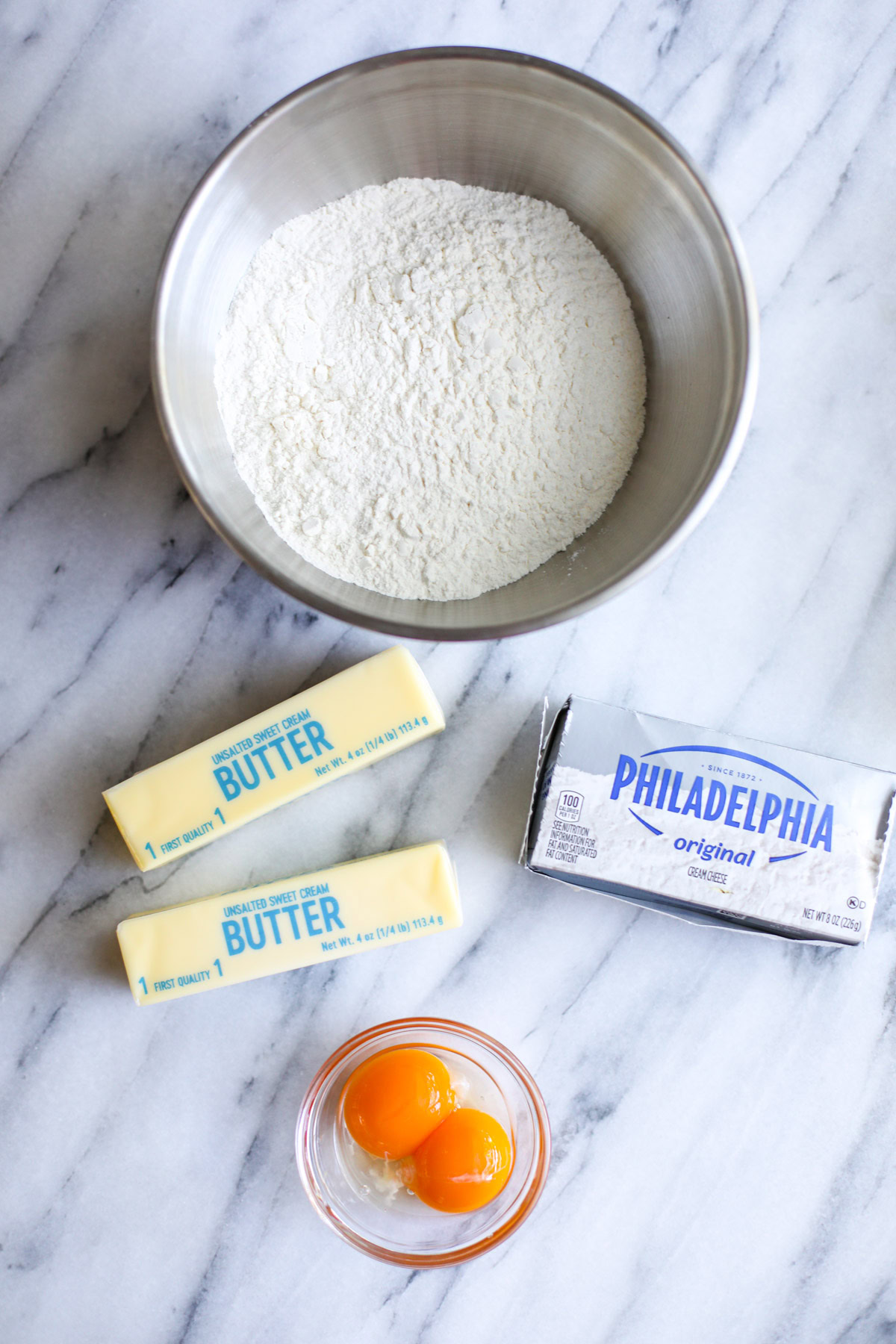 A stainless steel bowl of flour, 2 sticks of butter, a package of cream cheese, and 2 egg yolks in a glass bowl.