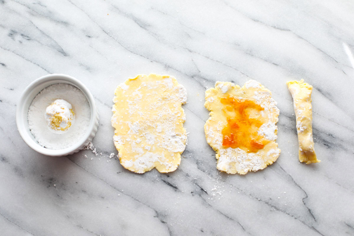 A small white bowl of powdered sugar, a rolled out cookie, a cookie with jam added down the center, and the cookie folded up into a log.