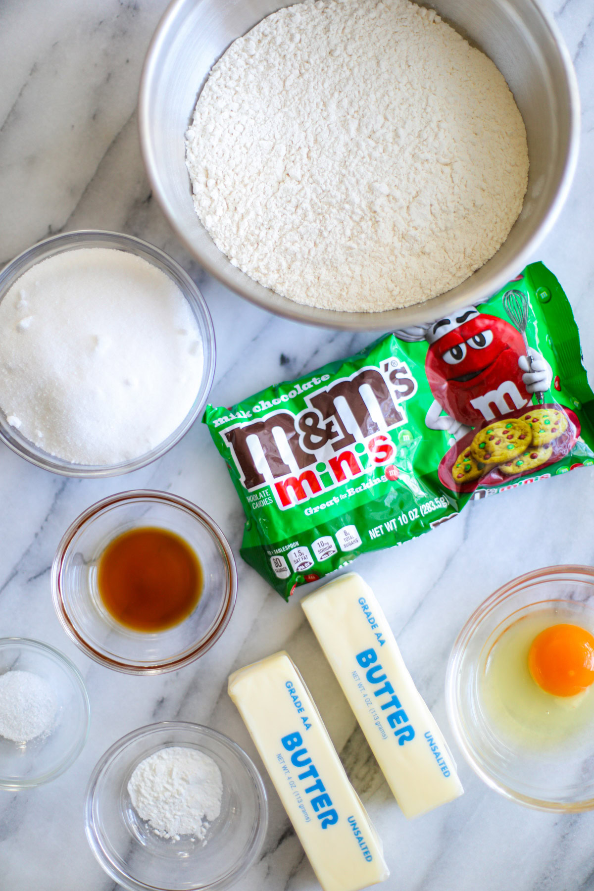 A stainless steel bowl of flour, a green and red bag of mini M&Ms, glass bowls of sugar, vanilla, egg, baking powder, salt, and 2 sticks of butter in the blue packaging.