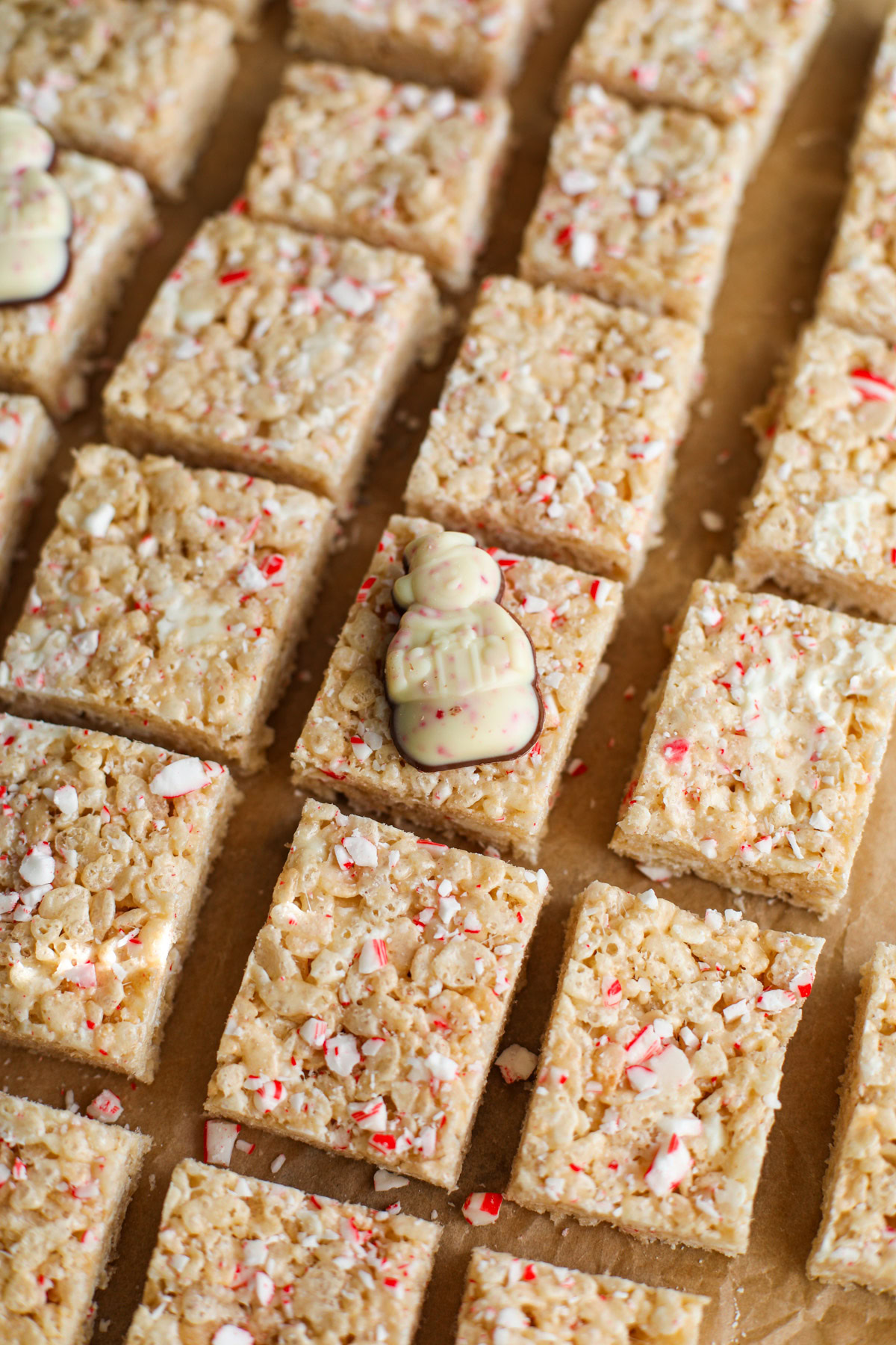 Rows of Rice Krispie treats on parchment paper topped with red and white pieces of candy cane and a peppermint bark shaped snowman candy.
