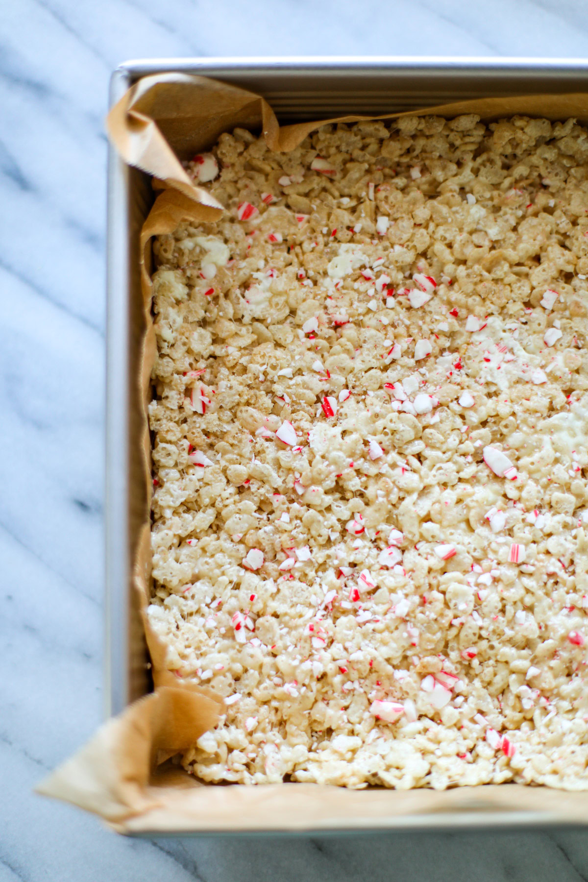 A metal sheet pan lined with brown parchment paper with tan, red, and white Rice Krispie treats left unsliced. 