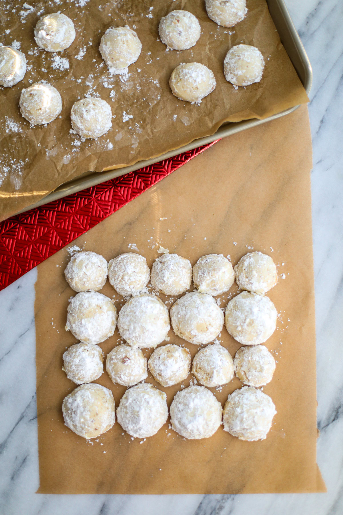 Small and large snowball cookies layered on brown parchment paper with a sheet pan of cookies in the back. 