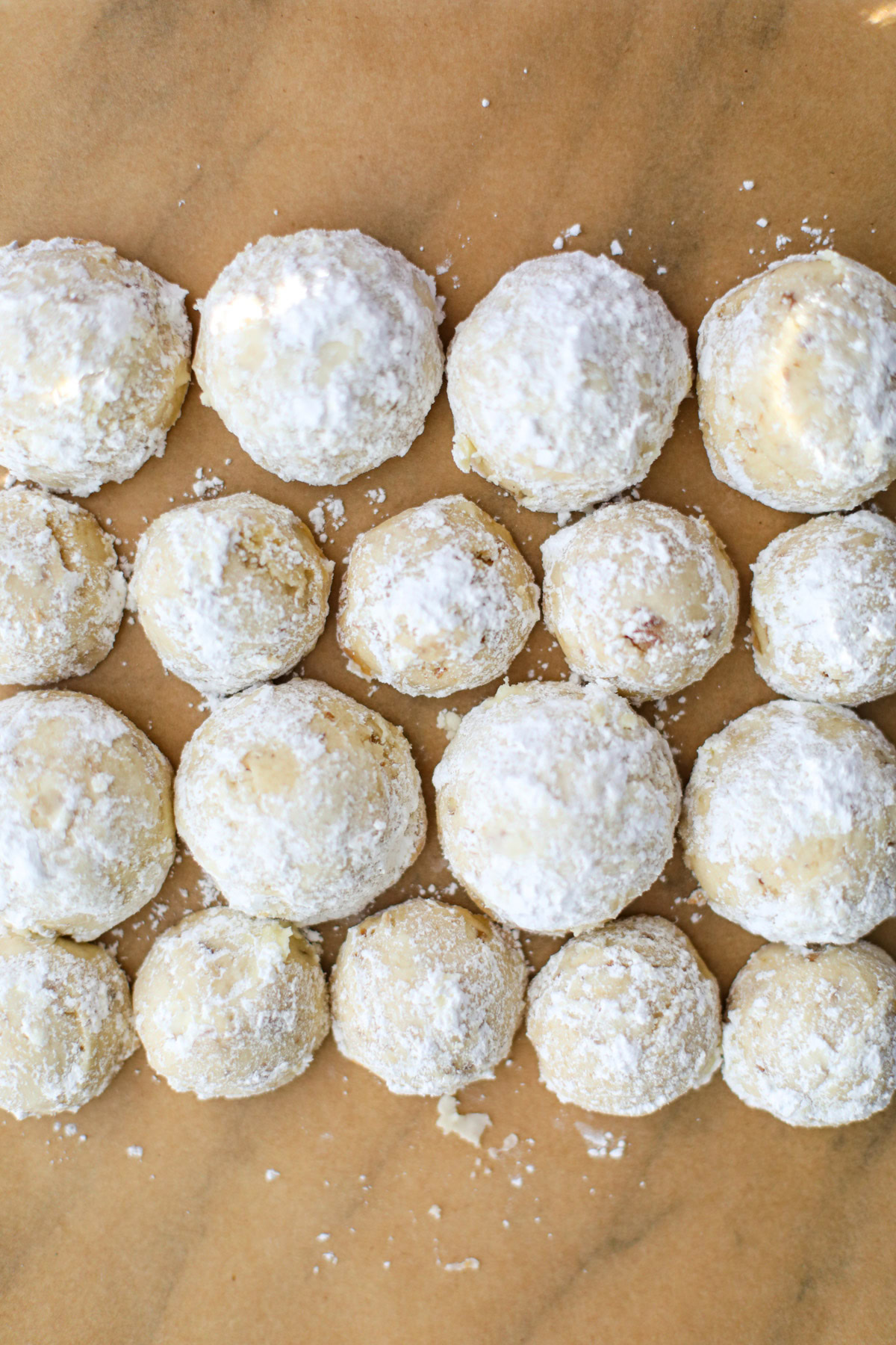 Four rows of small and large snowball cookies dusted with powdered sugar on brown parchment paper. 
