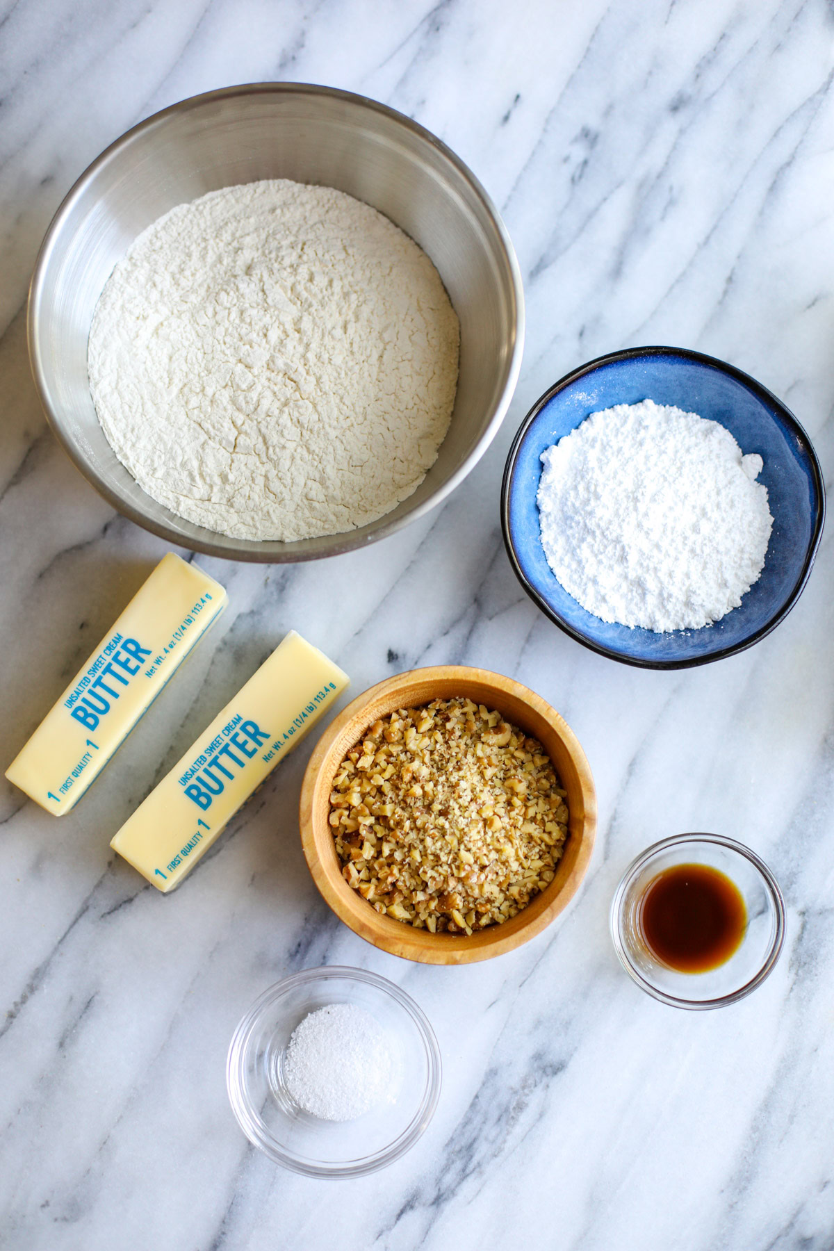 A bowl of flour, a blue bowl of powdered sugar, 2 sticks of butter, a wooden bowl of chopped walnuts, and two glass bowls of vanilla extract and kosher salt. 