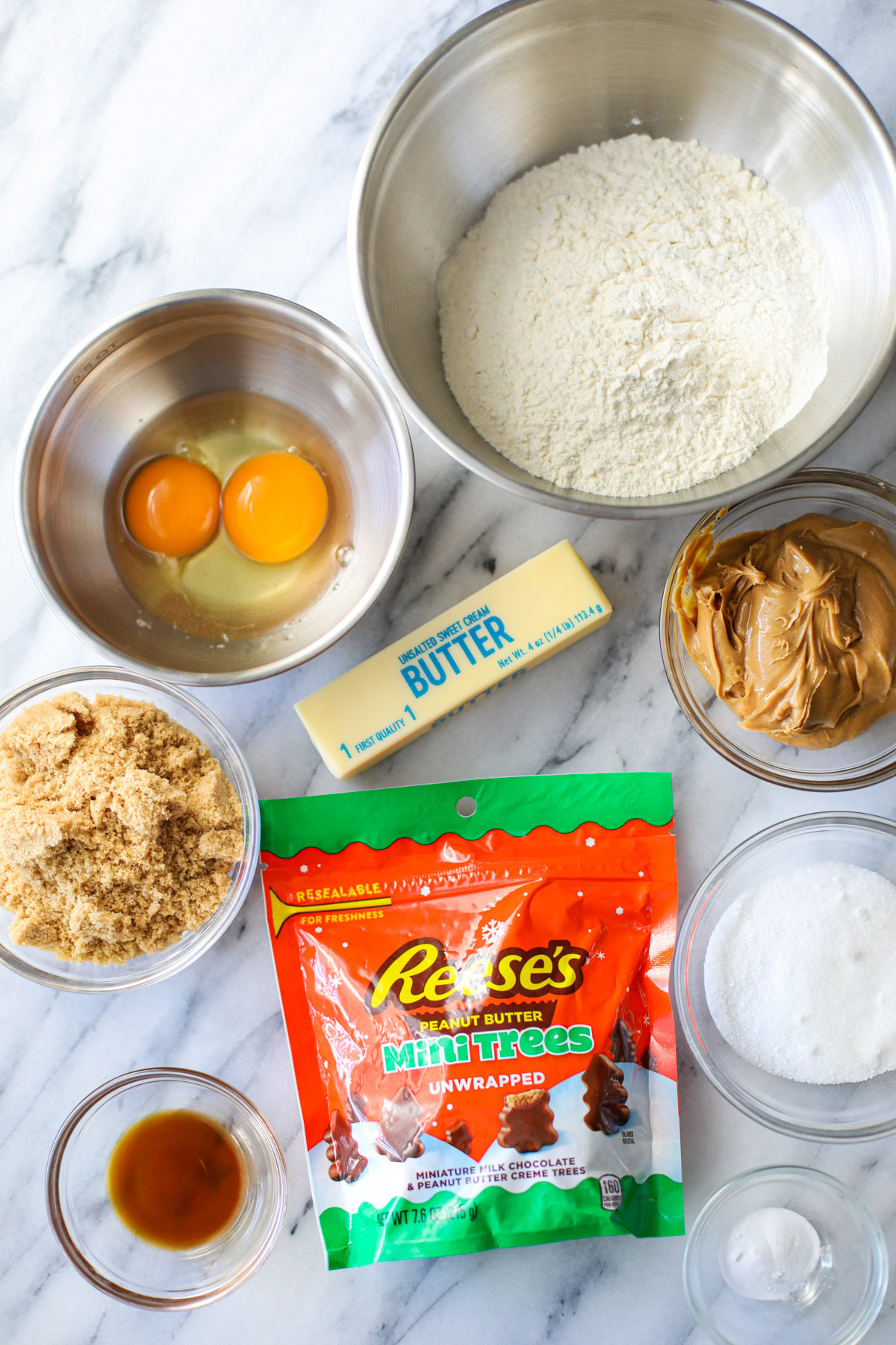 A stainless bowl of white flour, two cracked eggs, glass bowls of sugars, peanut butter, baking soda, vanilla, a wrapped stick of butter and a bag of Reese's Peanut Butter Mini Trees.