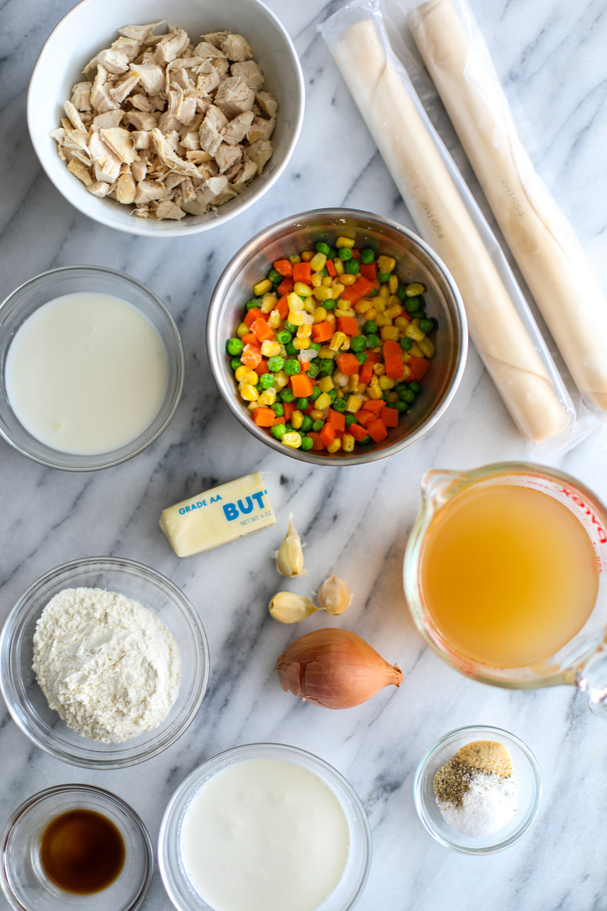 A white bowl of cubed chicken, two rolls of pie crust, glass bowls of cream, milk, chicken stock, seasonings, Worcestershire, flour, a metal bowl of veggies, a shallot, 3 cloves of garlic, and half a stick of wrapped butter. 