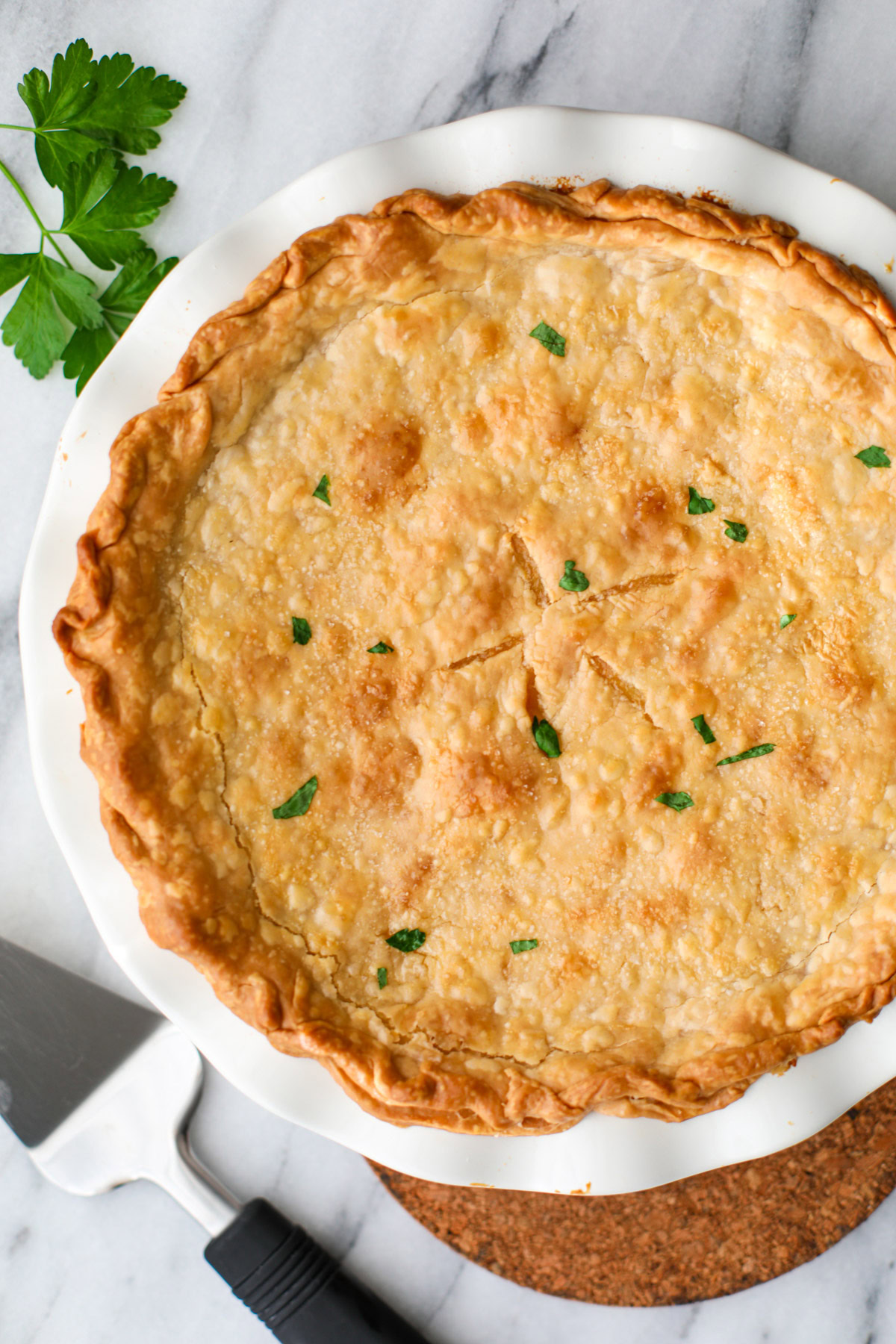 A full chicken pot pie in a white pie plate topped with pieces of parsley with a metal pie server on the sider. 