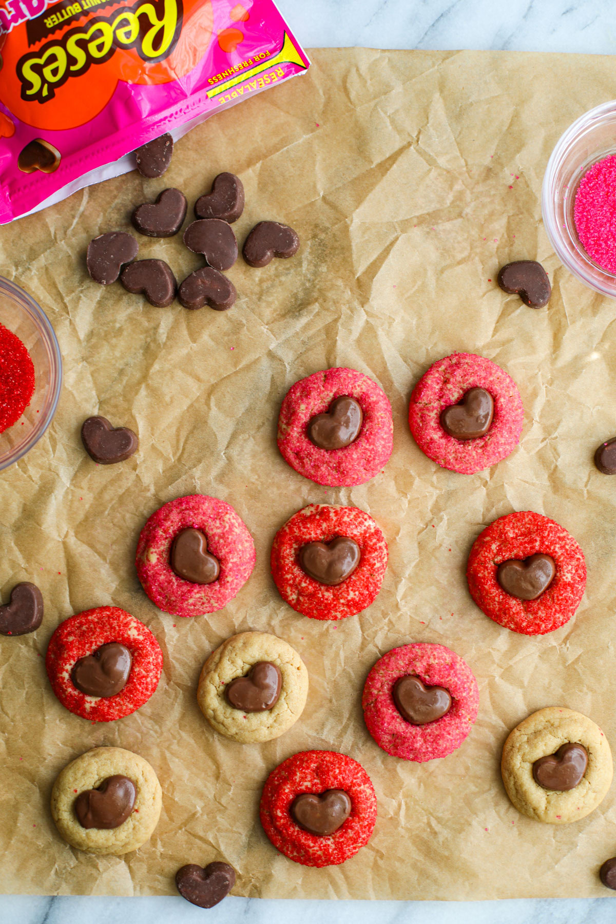 Red, pink, and plain peanut butter Cookies with mini Reese's hearts on crinkly brown parchment paper with bowls of red and pink sanding sugar. 