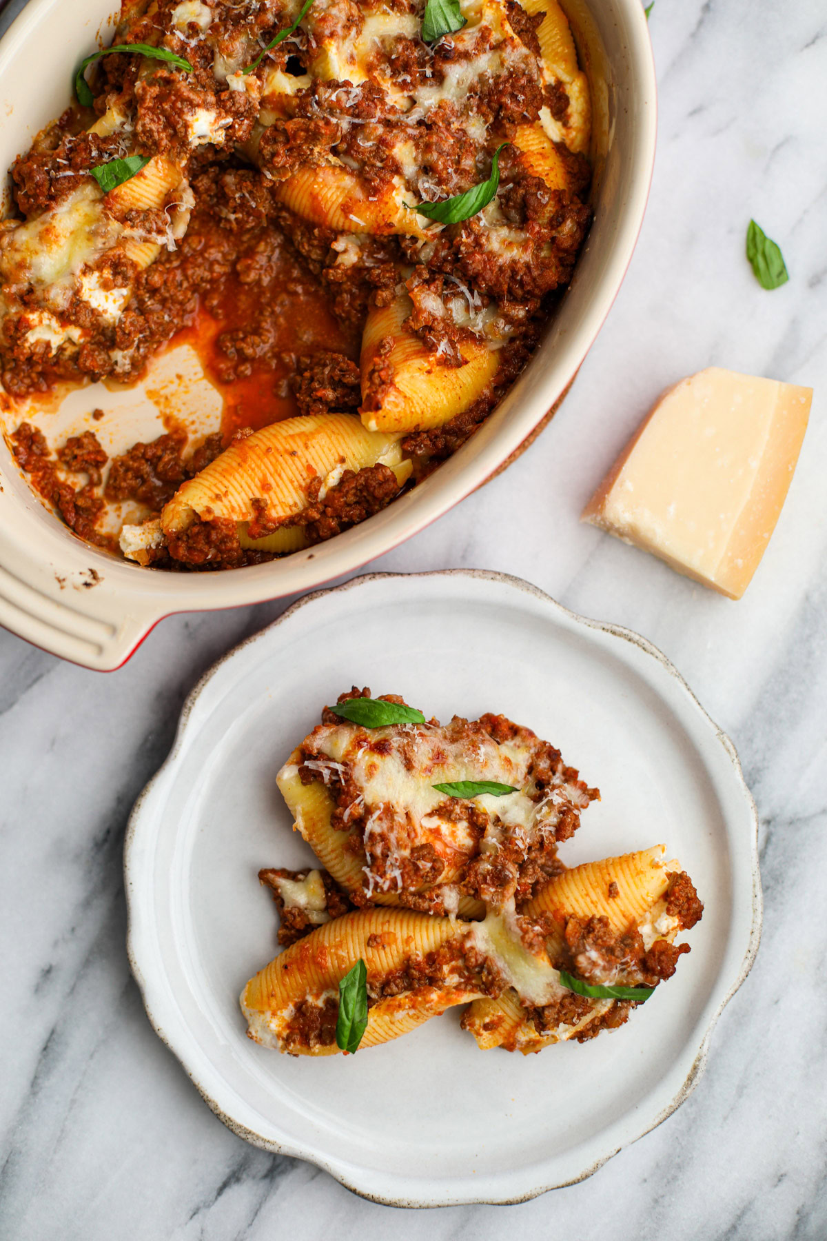 A round baking dish of saucy stuffed shells with a white plate with three shells topped with golden cheese and torn pieces of basil with a wedge of parmigiano reggiano in the background. 