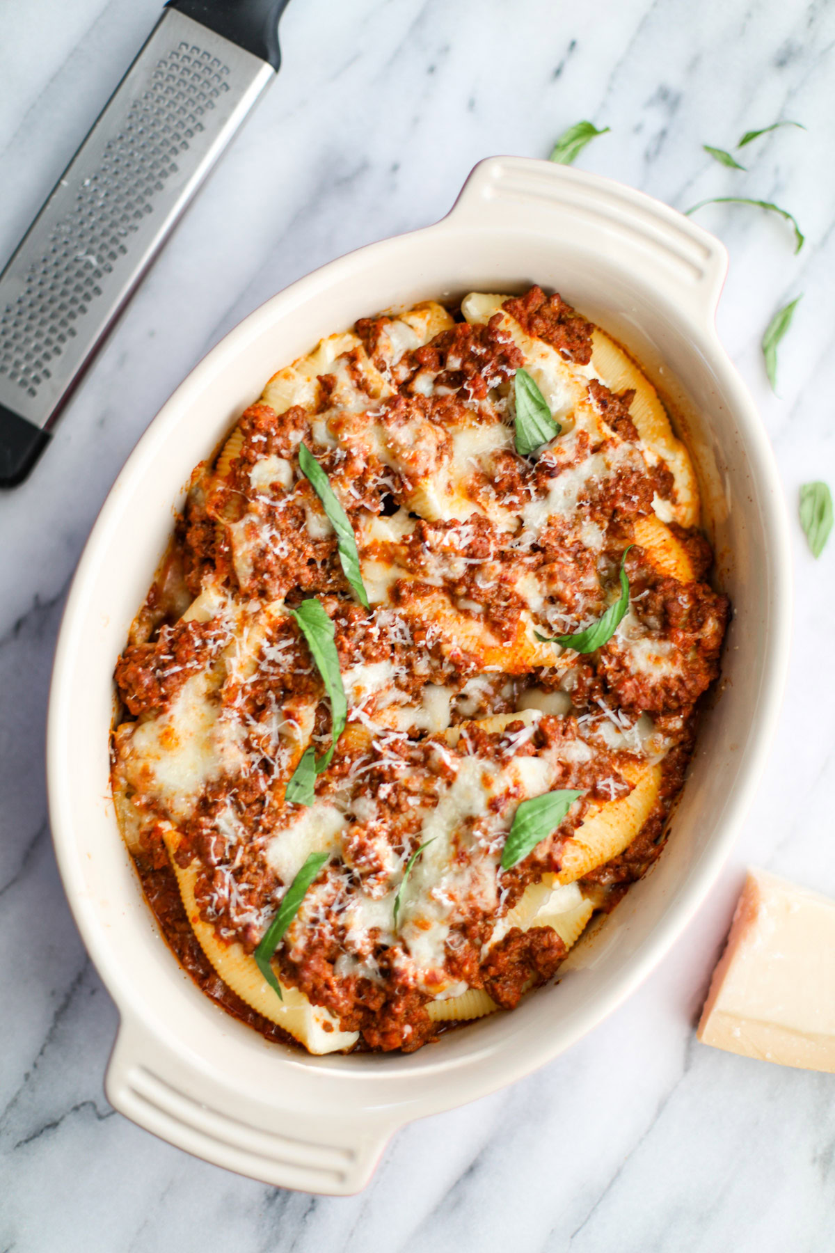 A finished oval baking dish of stuffed shells with torn leaves of basil and a microplane and wedge of cheese in the background.