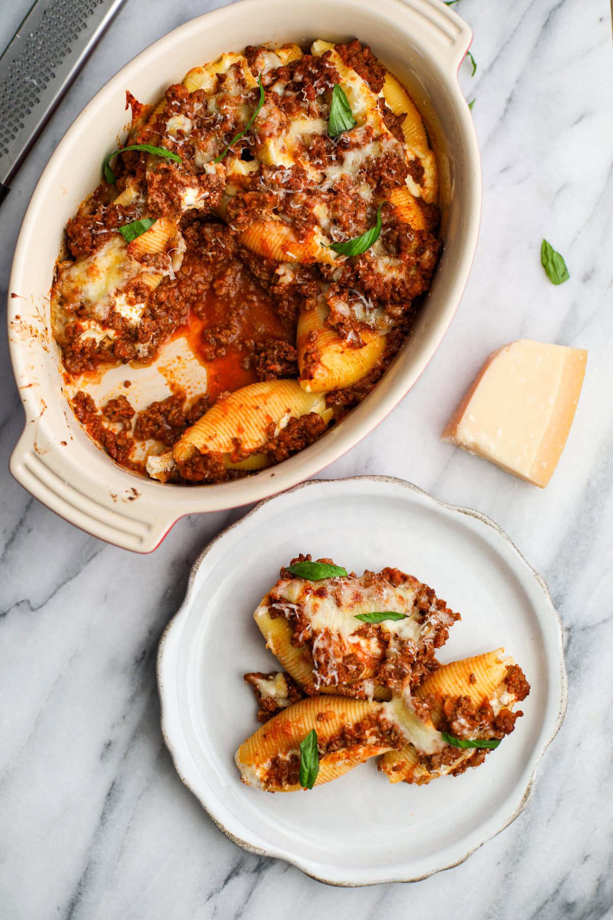 A round baking dish of saucy stuffed shells with a white plate with three shells topped with golden cheese and torn pieces of basil with a wedge of parmigiano reggiano in the background. 