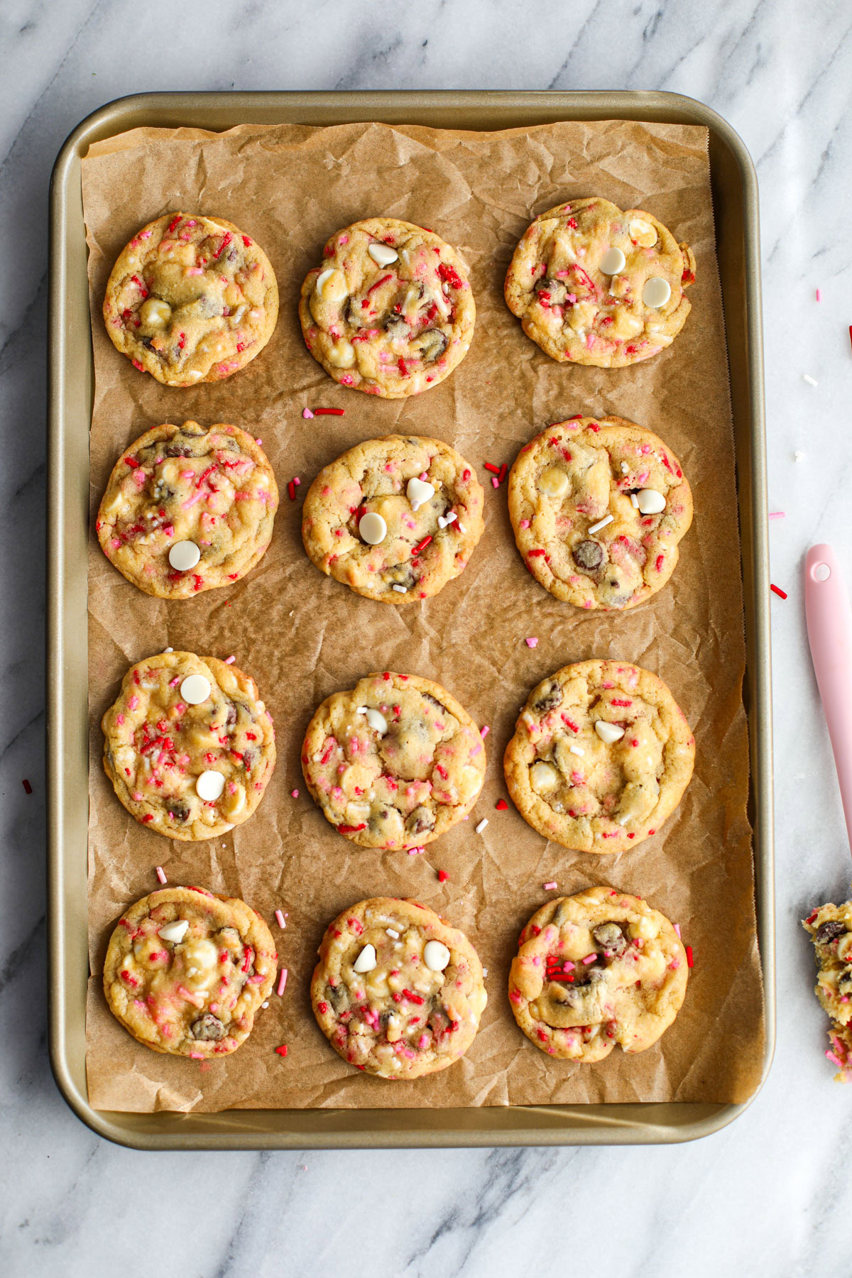 Twelve chocolate chip cookies with red and white sprinkles lined up on a gold cookie sheet.