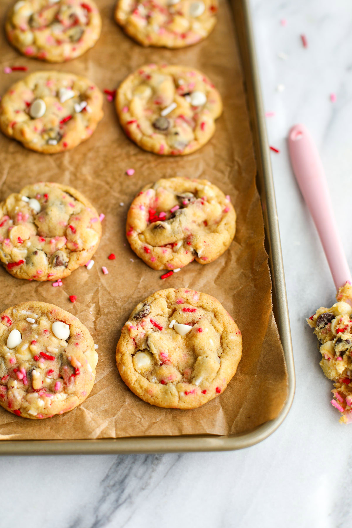 Chocolate chip cookies with red, pink, and white sprinkles lined up on a gold cookie sheet with a pink spatula covered in cookie dough.