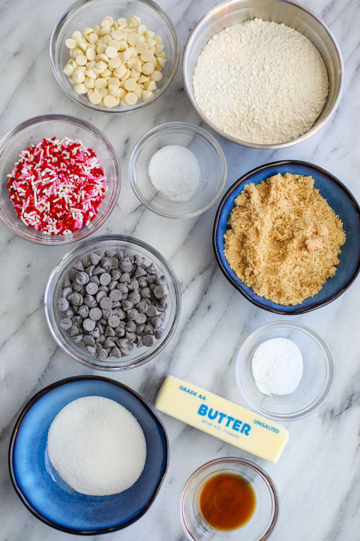 Clear bowls of chocolate chips, sprinkles, salt, baking soda, vanilla, and blue bowls of light brown and white sugar with a stick of butter and a stainless bowl of flour.