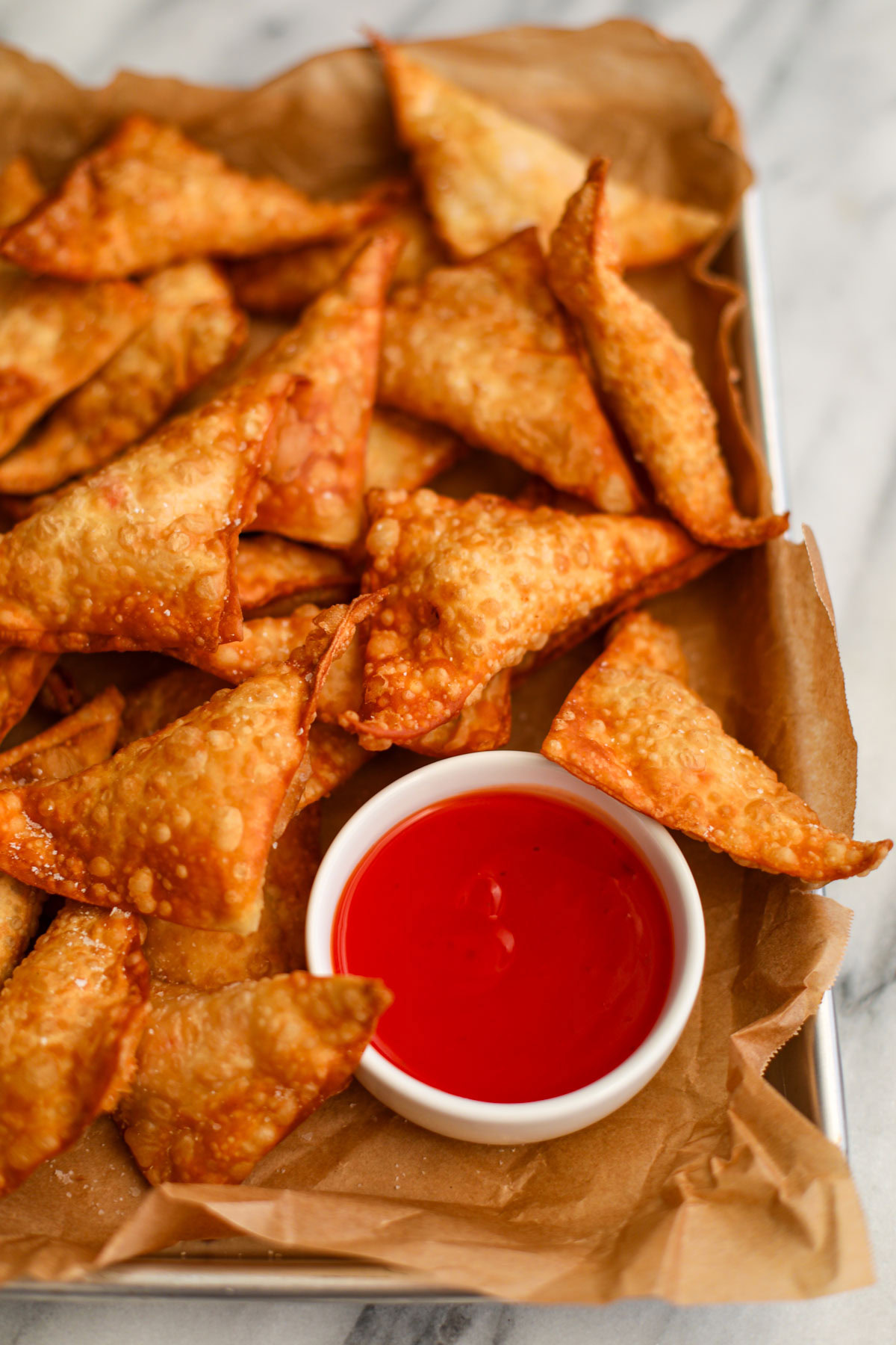 A metal cookie sheet lined with parchment paper topped with a dozen fried crab rangoon and a white bowl of bright red sweet and sour sauce.