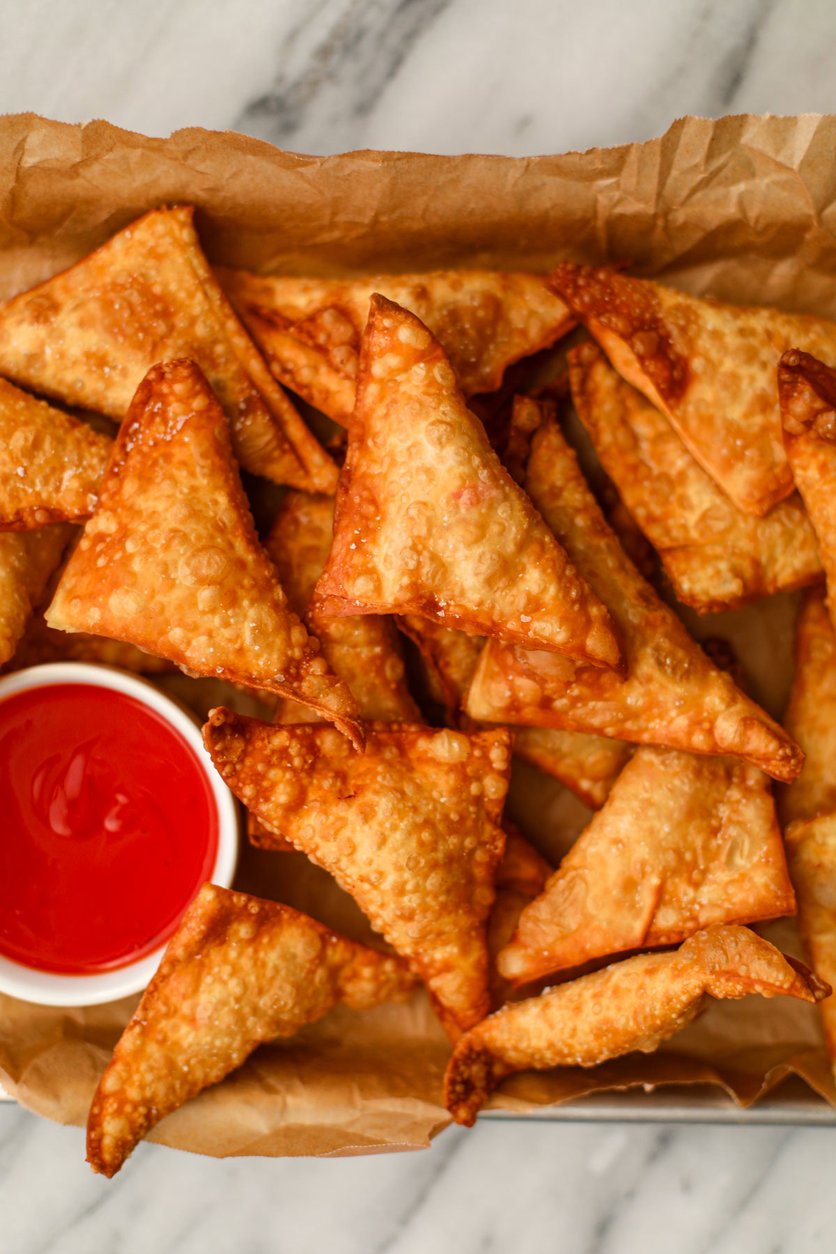 Classic crab rangoon and a white bowl of bright red sweet and sour sauce on a white counter.
