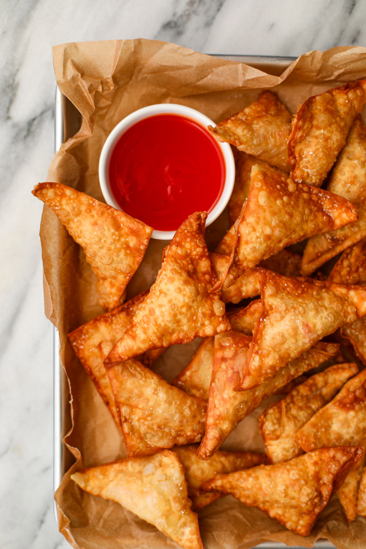 A metal cookie sheet lined with parchment paper topped with a dozen fried crab rangoon and a white bowl of bright red sweet and sour sauce.