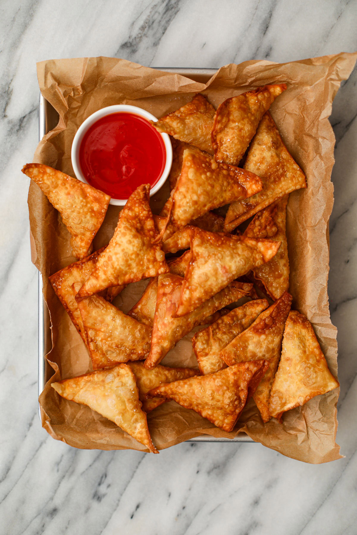 A metal cookie sheet lined with parchment paper topped with a dozen fried crab rangoon and a white bowl of bright red sweet and sour sauce sitting on a white marble counter.
