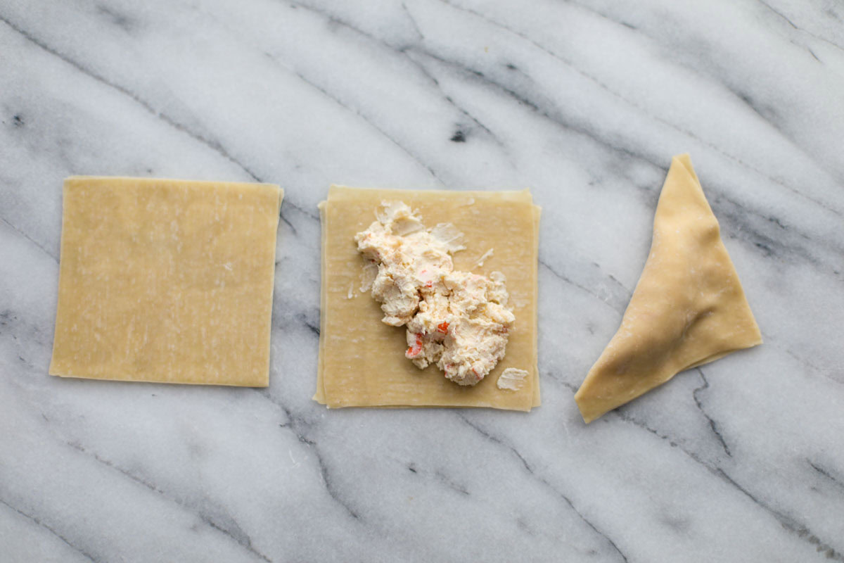 A pile of wonton wrappers, a wrapper with crab rangoon filling smeared down the center, and the wonton wrapper folded over the filling in half. 
