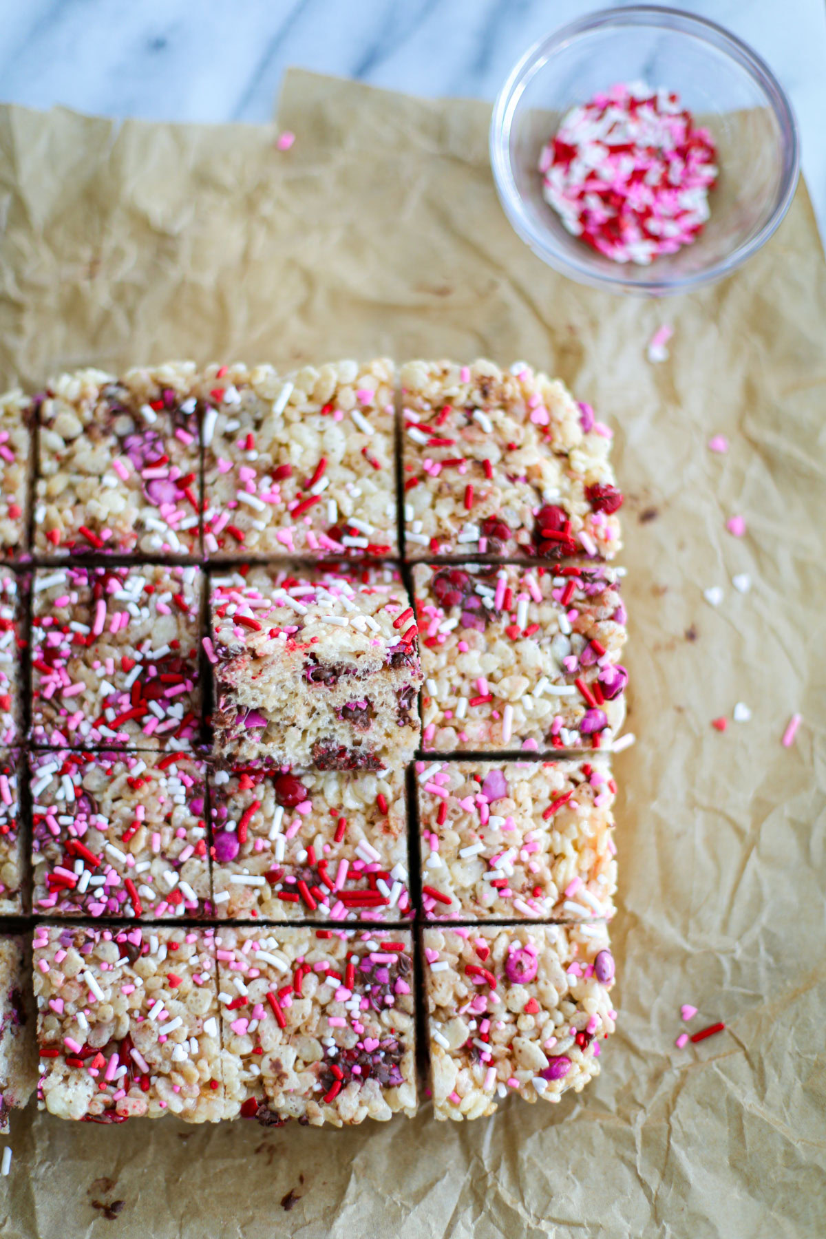 Pink, red, and white sprinkled Valentine's Mini M&M Rice Krispie treats cut into small squares lined up on crinkled brown parchment paper.