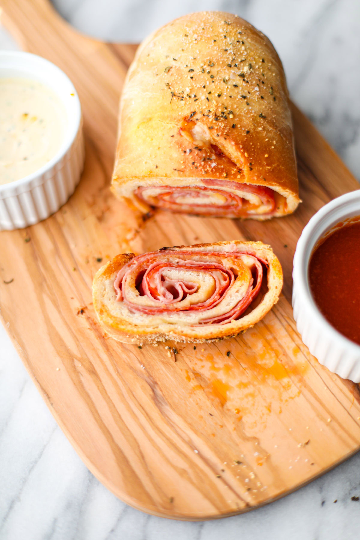 One slice of stromboli on an oblong olive wood cutting board with a white ramekin of Italian dressing and a white ramekin of marinara.