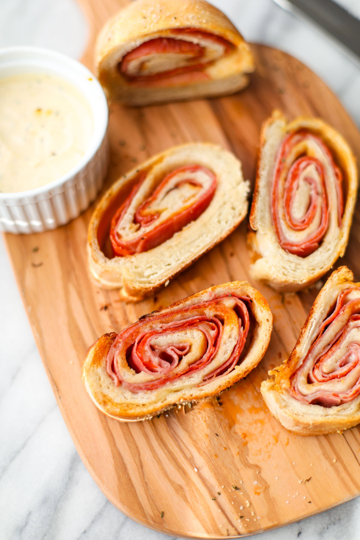 Four slices of stromboli on an oblong olive wood cutting board with a white ramekin of Italian dressing.