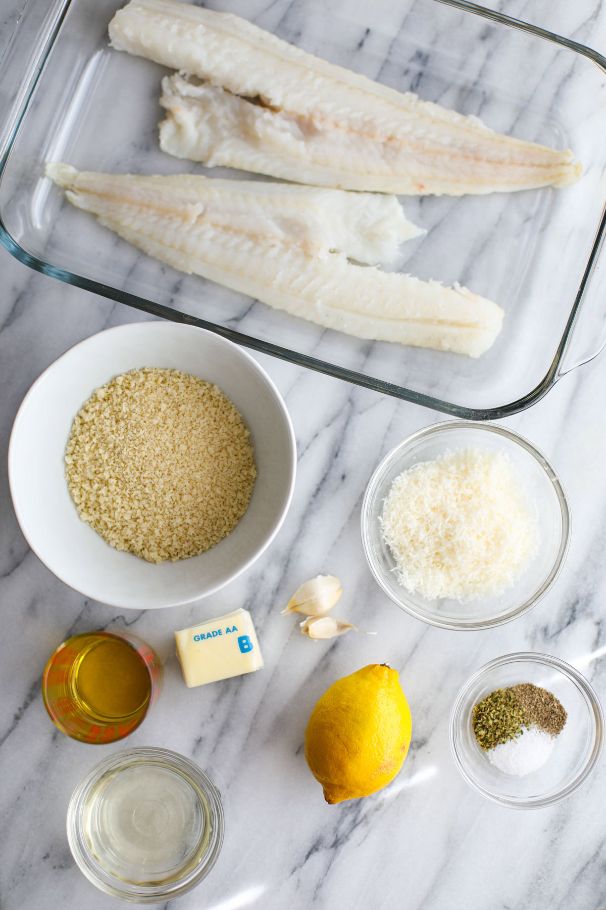 All the ingredients to make a cod oreganata including a glass dish of two cod loins, a white bowl of breadcrumbs, glass bowls of cheese, seasonings, olive oil, and white wine with a whole lemon and 2 garlic cloves. 