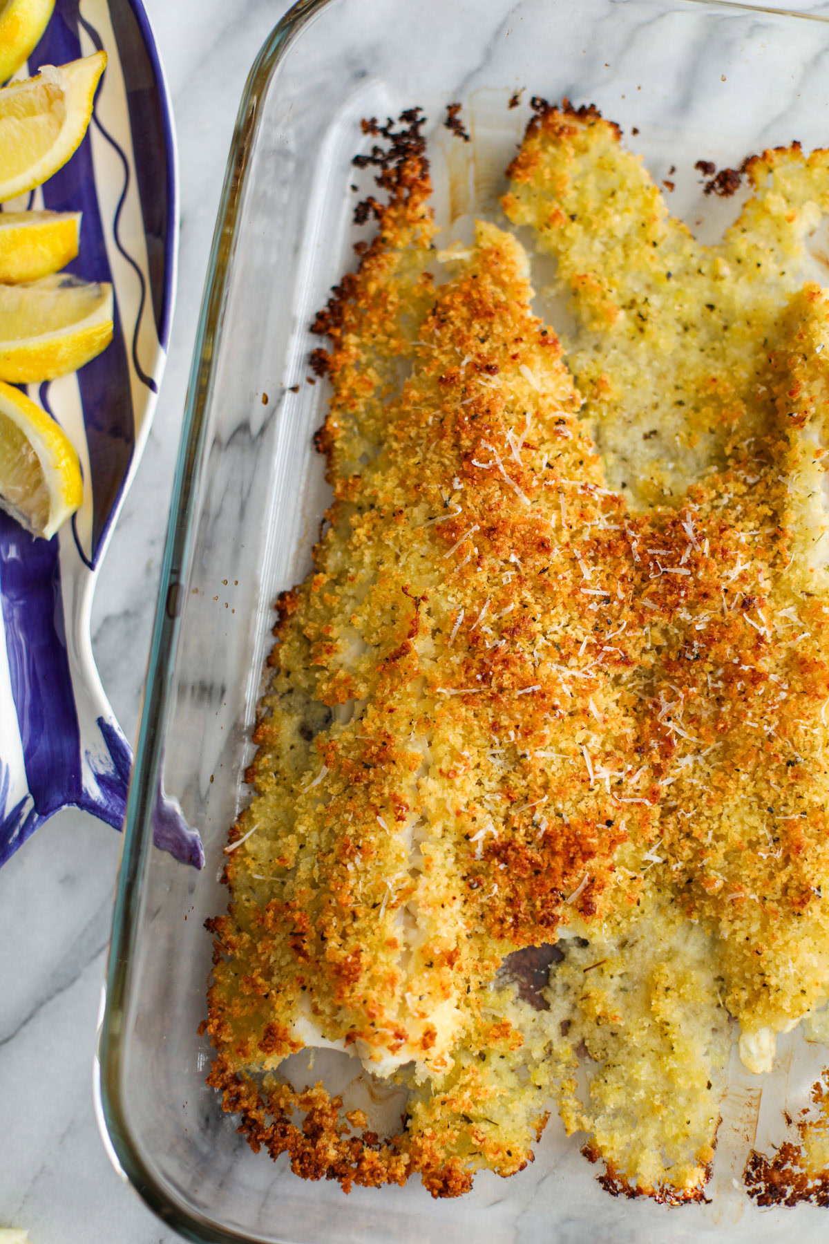 A glass baking dish with cod oreganata topped with a crispy golden breadcrumb mixture with a blue fish dish topped with slices of lemons in the back.
