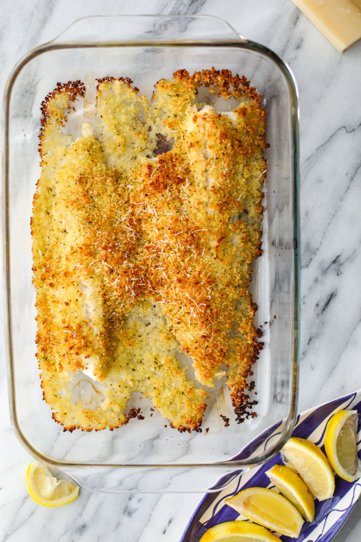 A glass baking dish with fish topped with a crispy golden breadcrumb mixture with a blue fish dish topped with slices of lemons and a large wedge of parmigiano.