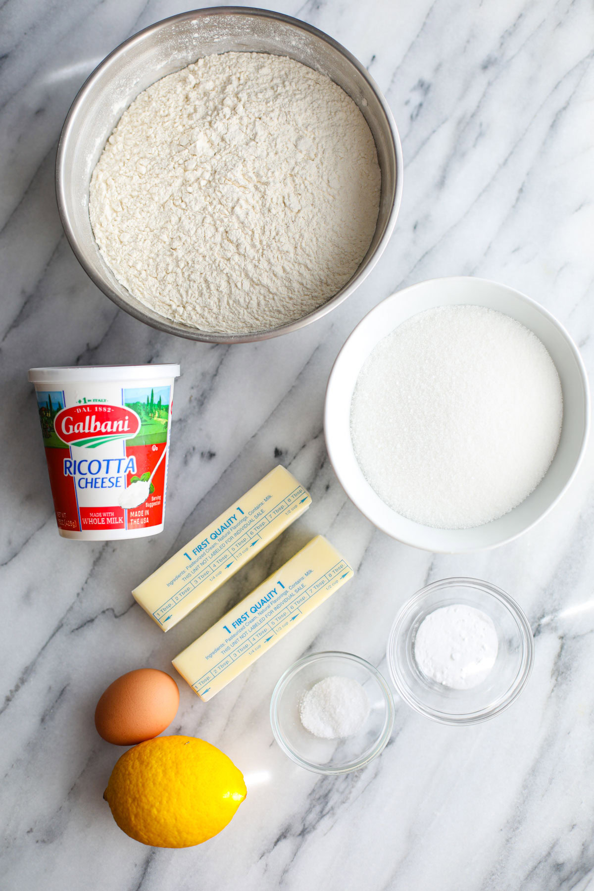 A stainless steel bowl of flour, a white bowl of sugar, a container of galbani ricotta cheese, 2 sticks of butter, 1 egg, a whole lemon, and small glass bowls of salt and baking soda.