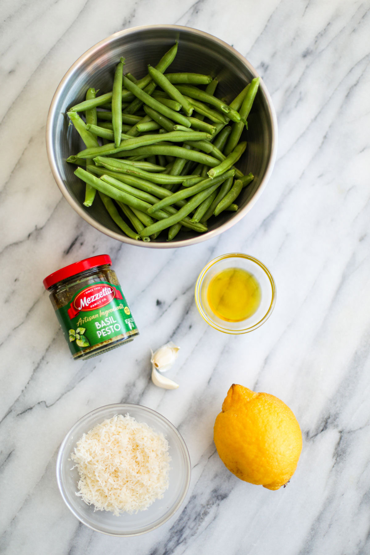 A stainless steel bowl of green beans, a clear bowl of olive oil, a jar of Mezzetta pesto, a whole lemon, 2 cloves of garlic and a clear bowl of grated parmigiano.