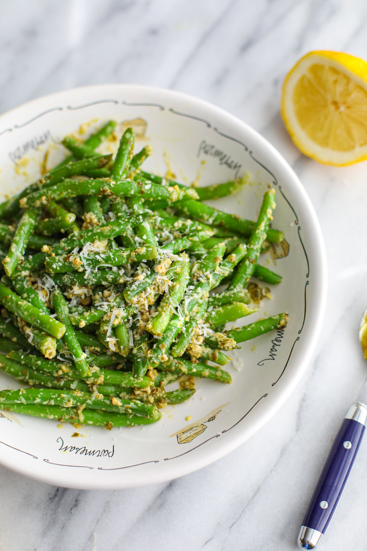 Green beans tossed with pesto in a shallow white bowl with half a lemon and a blue spoon off to the side.