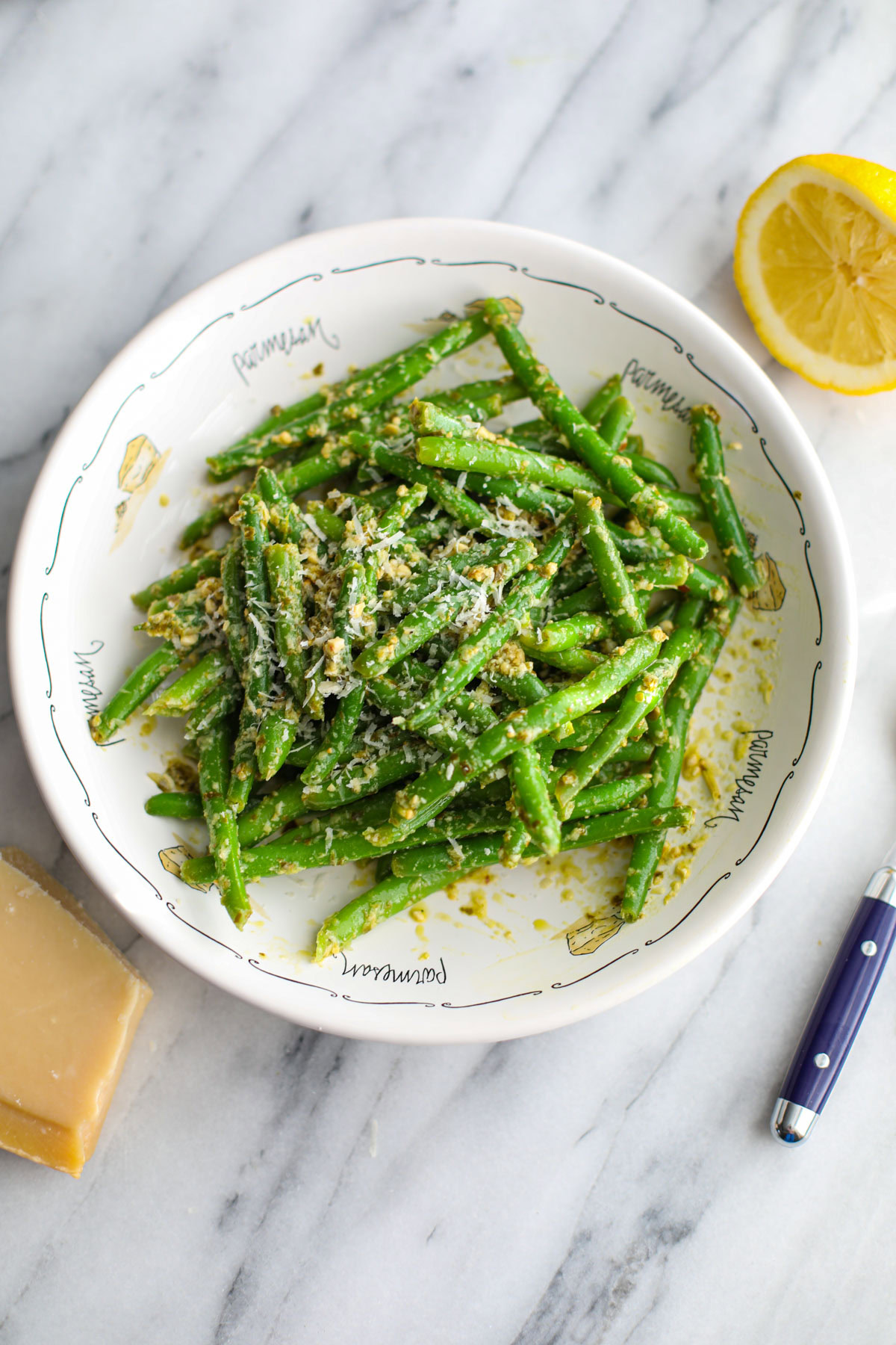 Green beans tossed with pesto in a shallow white bowl with half a lemon, a blue spoon, and a wedge of parmesan off to the side.