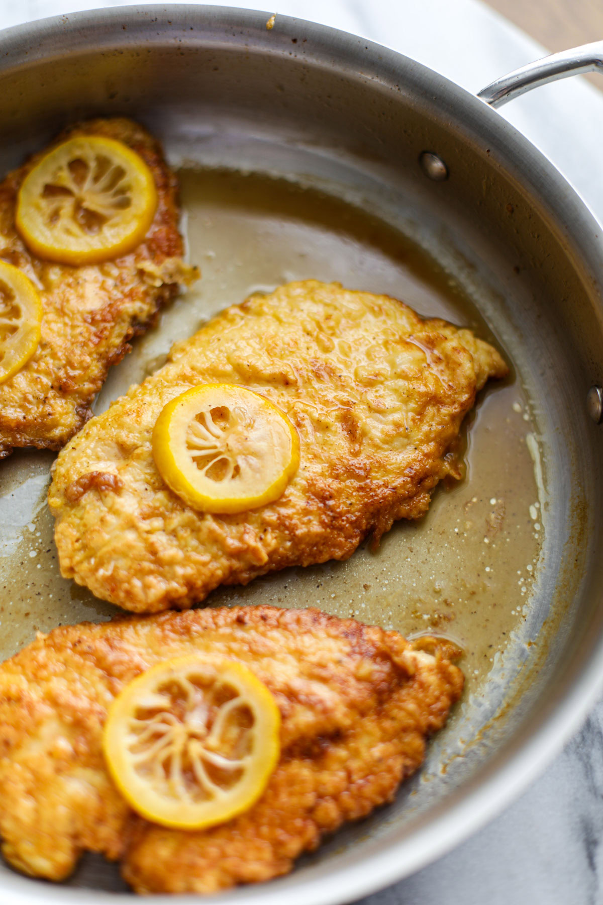 Three pieces of chicken francese topped with slices of lemon in a stainless steel skillet. 