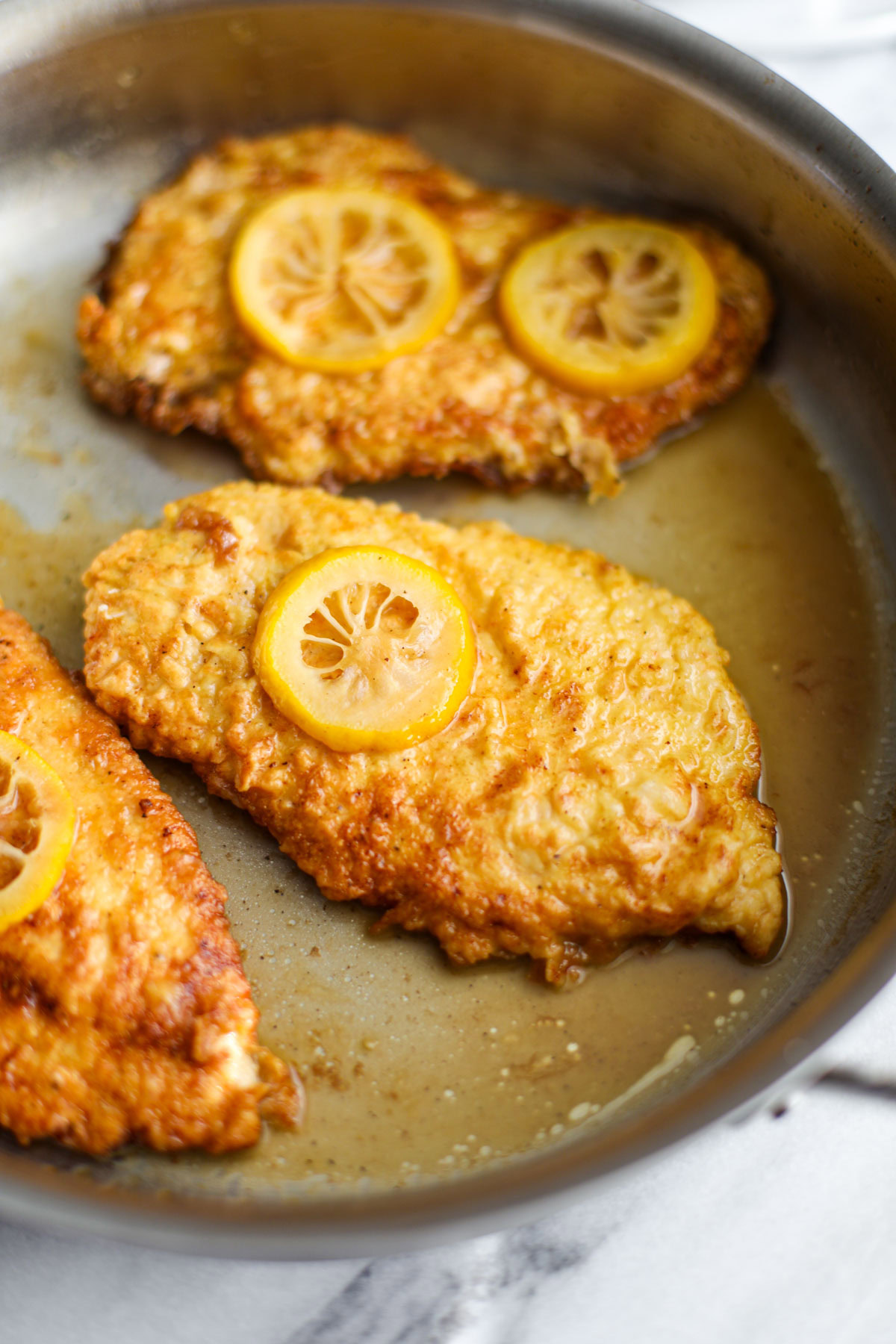 Three pieces of chicken francese topped with slices of lemon in a stainless steel skillet. 