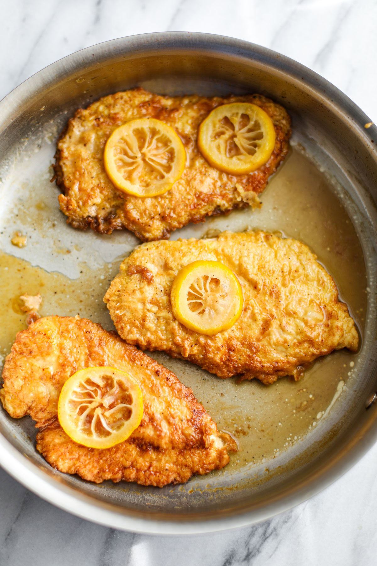 Three pieces of chicken francese topped with slices of lemon in a stainless steel skillet. 
