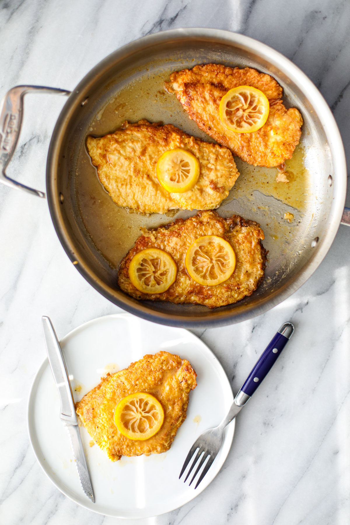 Three pieces of chicken francese topped with slices of lemon in a stainless steel skillet with a white plate of chicken and a blue fork and stainless steel knife. 