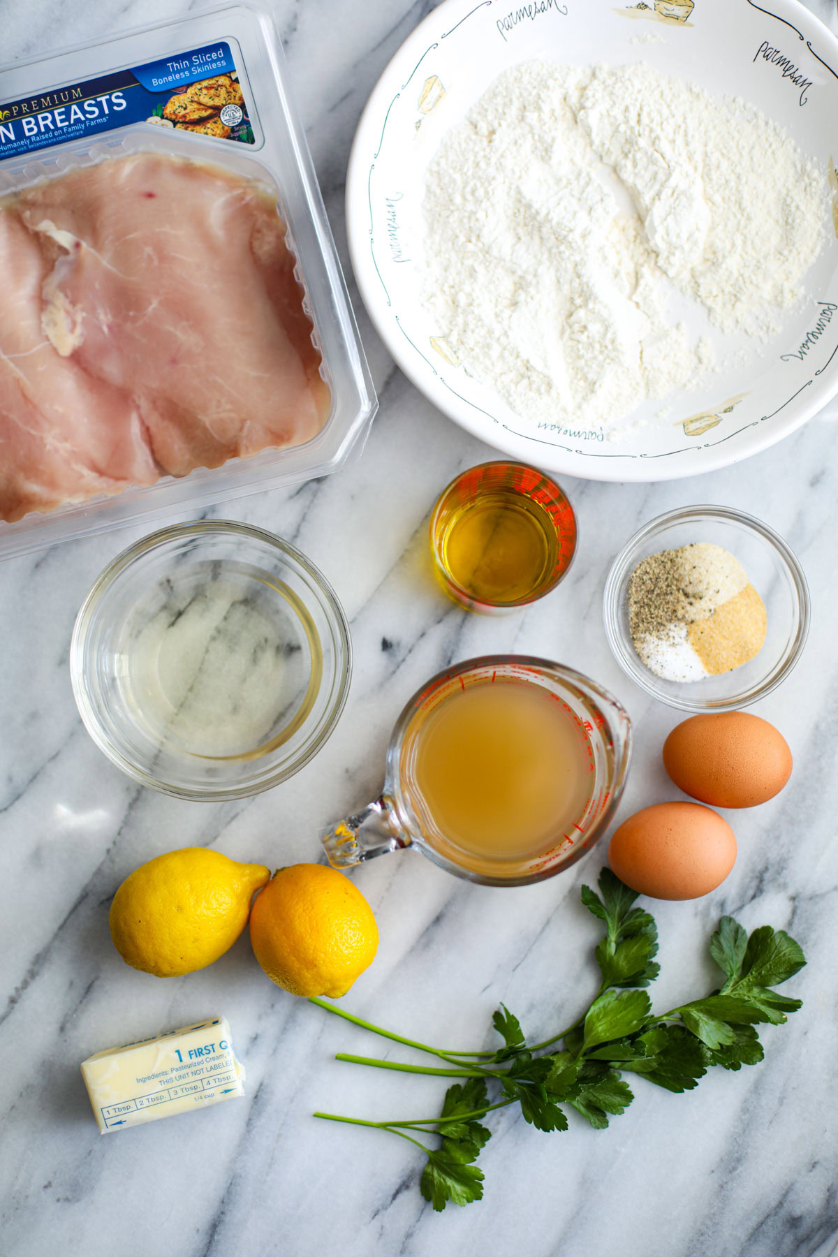 A bowl of flour, a package of chicken breasts, glass bowls of wine and seasoning, a glass measuring cup of chicken stock, 2 eggs, 2 lemons, a stick of butter and two stalks of fresh parsley. 