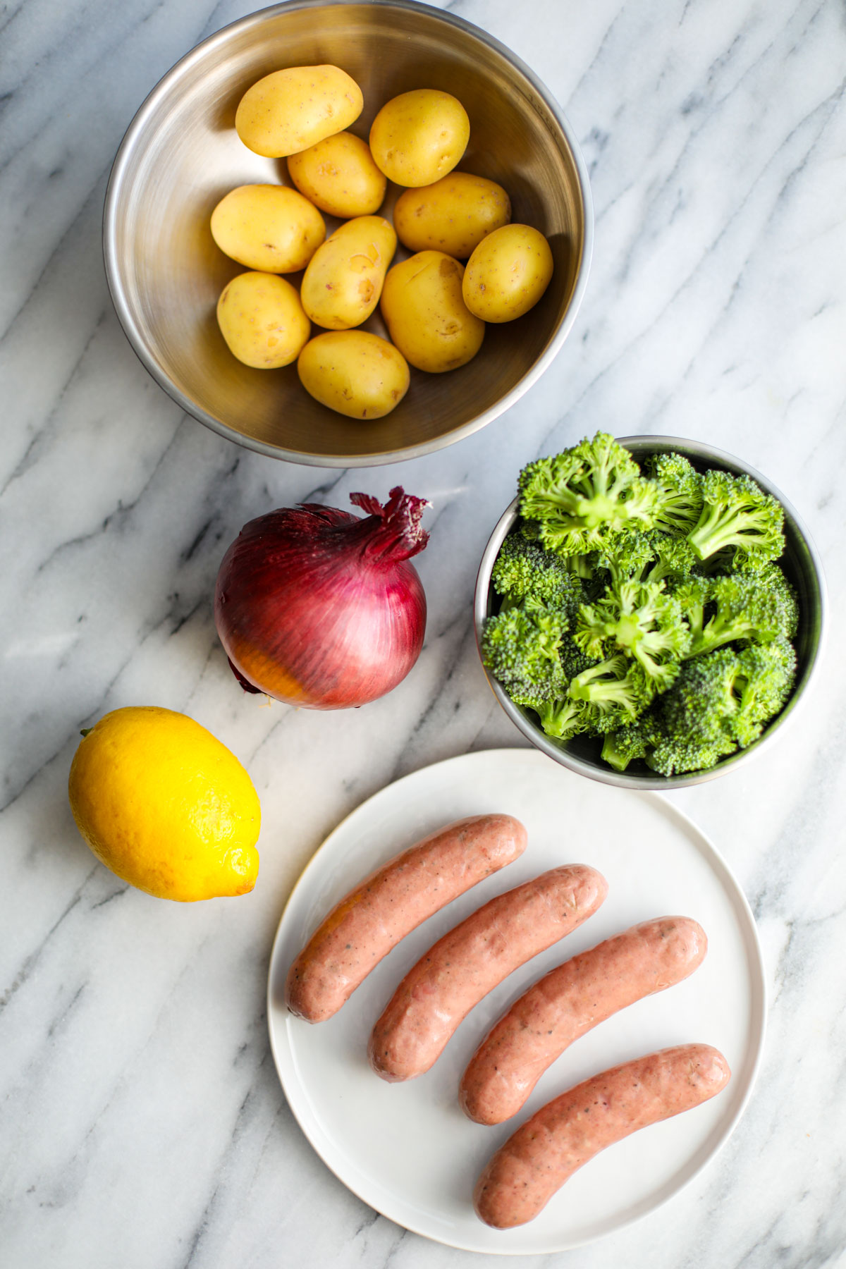 A stainless steel bowl of baby gold potatoes, a bowl of broccoli florets, a red onion, a lemon, and 4 chicken sausages on a white plate. 
