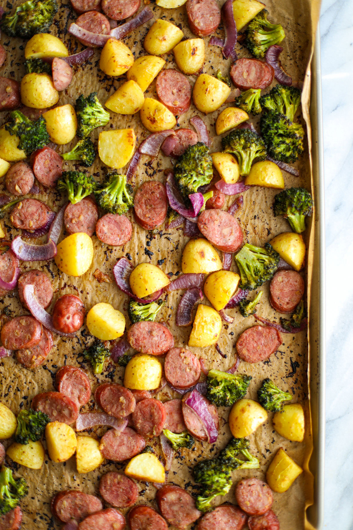 A sheet pan lined with parchment with sliced chicken sausage, baby gold potatoes, broccoli florets, and sliced red onion.